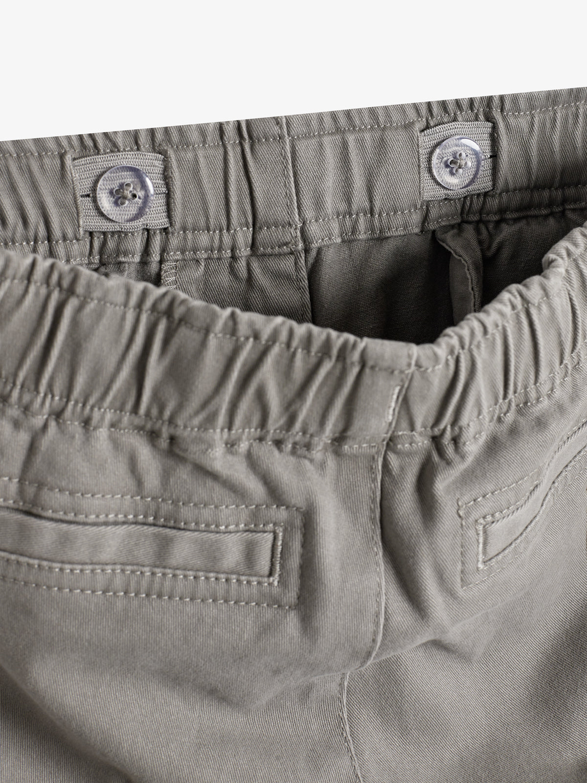 Gray elastic waistband with adjustable buttons is shown on gray fabric pants, featuring two rear welt pockets. The focus is on the waistband and stitching detail.