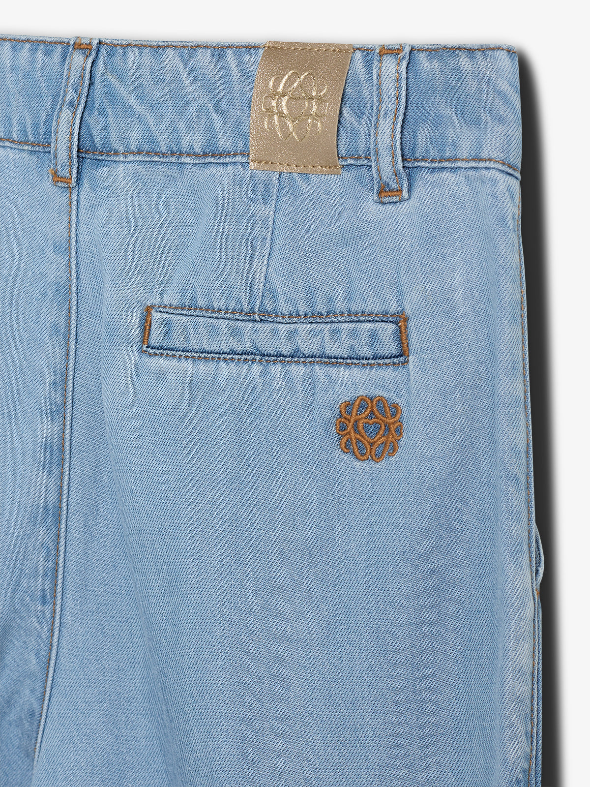 Light blue denim features a small pocket and belt loops, embroidered with an intricate brown logo. The background is plain, highlighting the fabric’s texture and stitching details.