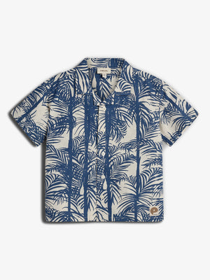 A short-sleeve, button-up shirt features a blue palm tree pattern on a beige background. It includes a small brown patch logo at the lower right corner.