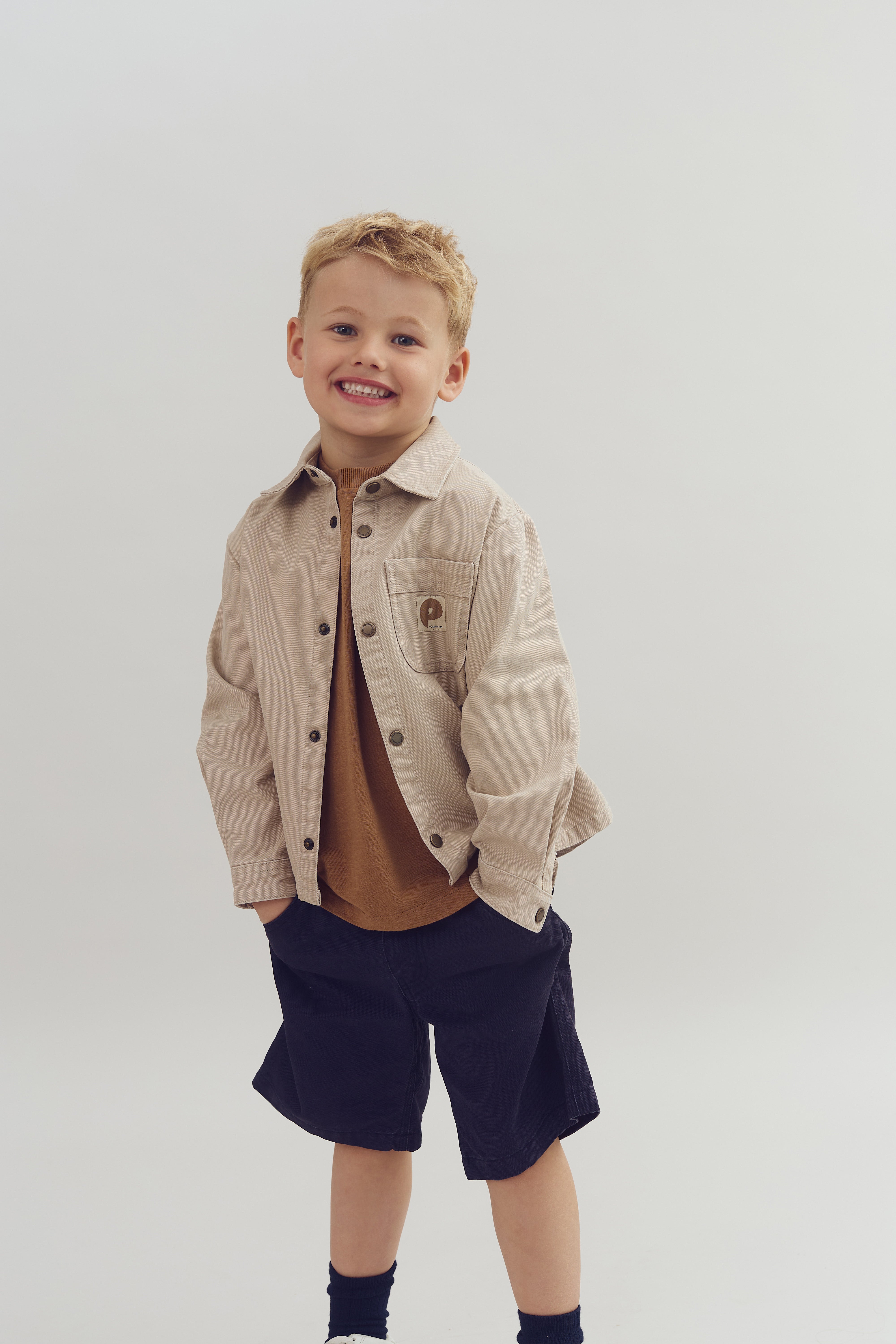 Child stands smiling, wearing a beige jacket with a pocket logo and dark shorts, hands in pockets, against a plain light background.