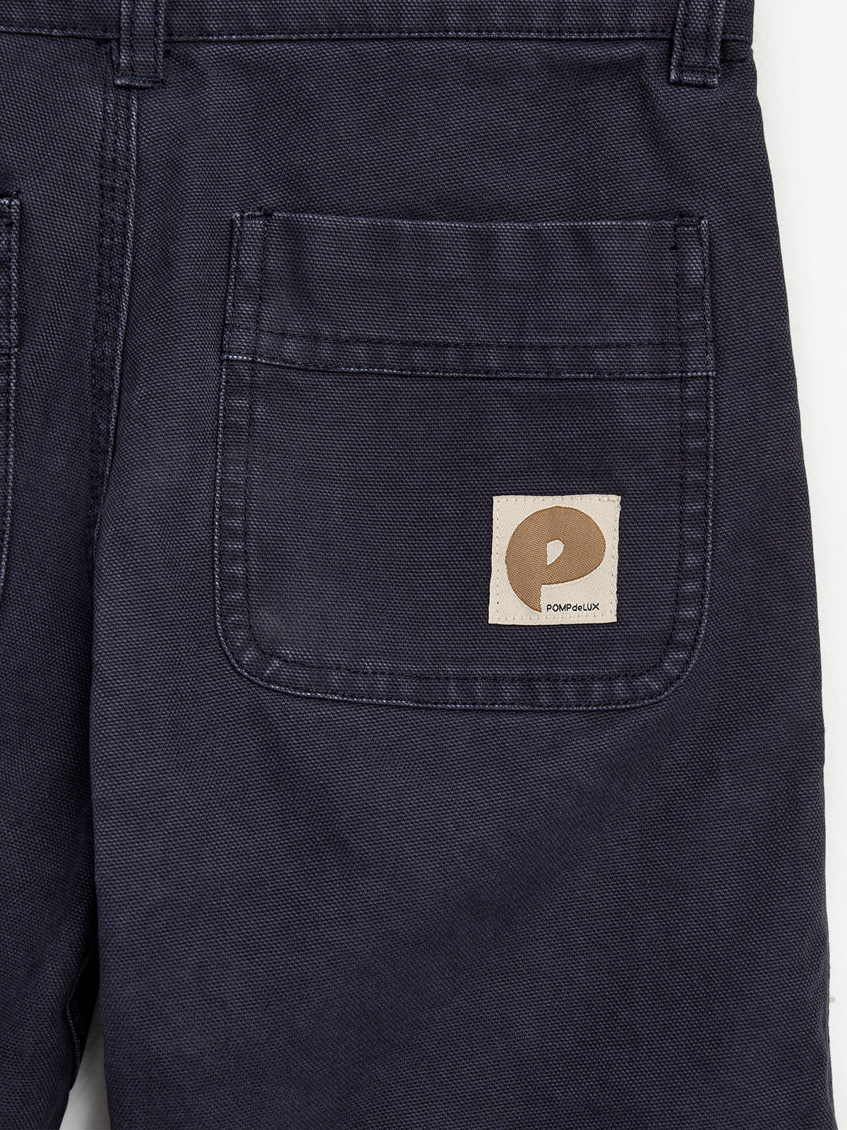Dark blue fabric with a large pocket featuring a beige tag displaying a stylized "P" and text "POMPdeLUX”; part of a garment, possibly pants, in a plain setting.
