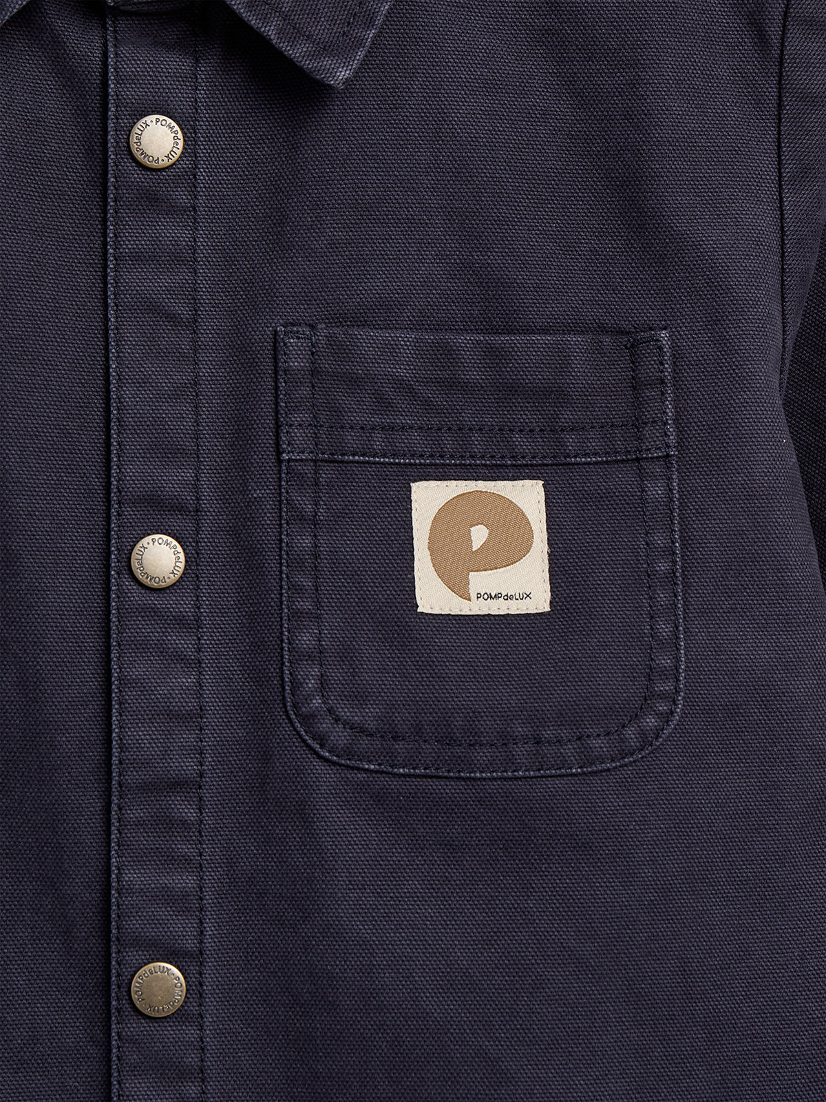 A navy blue shirt features a front pocket with a sewn beige label displaying a "P" and "POMPdeLUX." Silver buttons are embossed with "POMPdeLUX" branding along the placket.