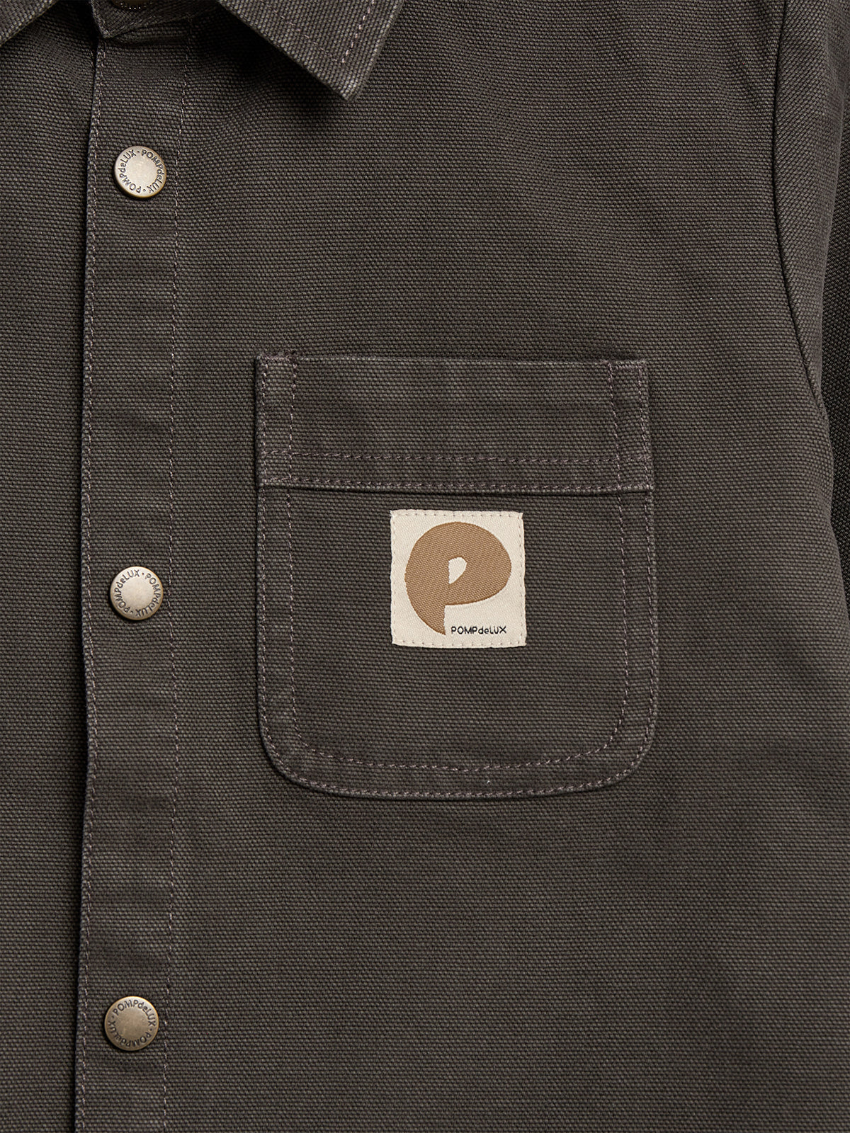 A dark fabric shirt with metal buttons and a chest pocket featuring a beige logo patch reading "POMPdeLUX" in a textured material setting.