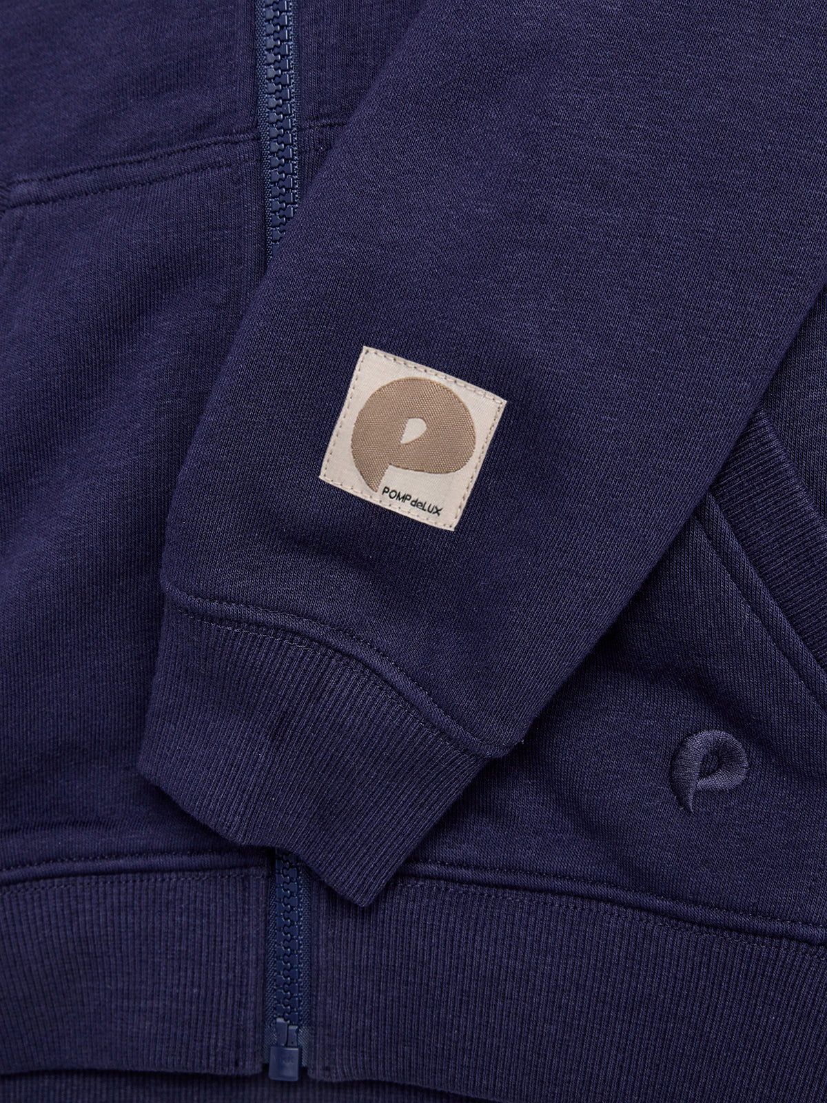 A dark blue hoodie features a visible zipper and a small brown fabric tag with a "P" logo and "POMPdeLUX" text. The sleeve rests on the body of the hoodie.