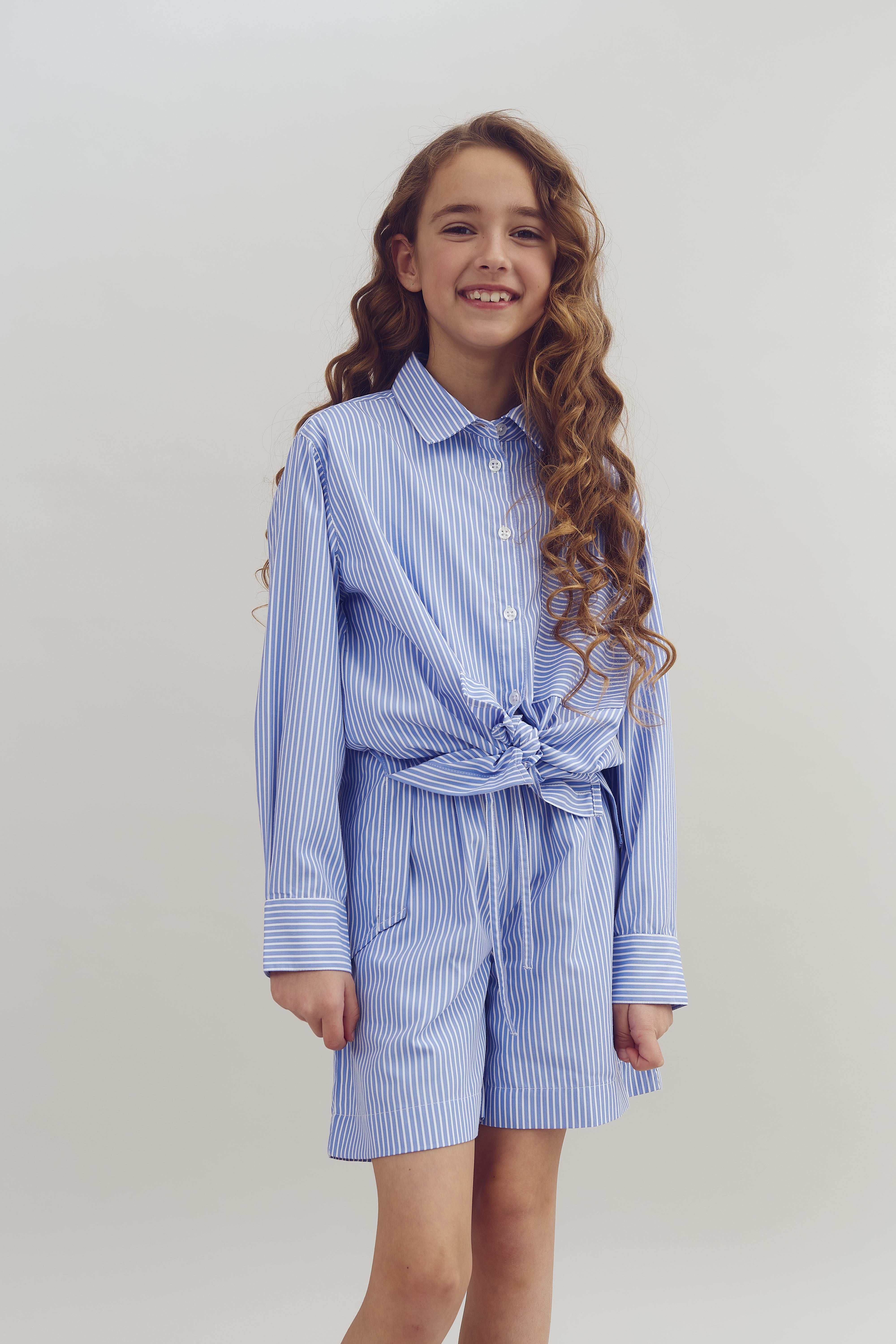A young girl with long, curly hair smiles while standing. She wears a blue and white striped shirt and shorts, tied at the waist, against a plain, light background.