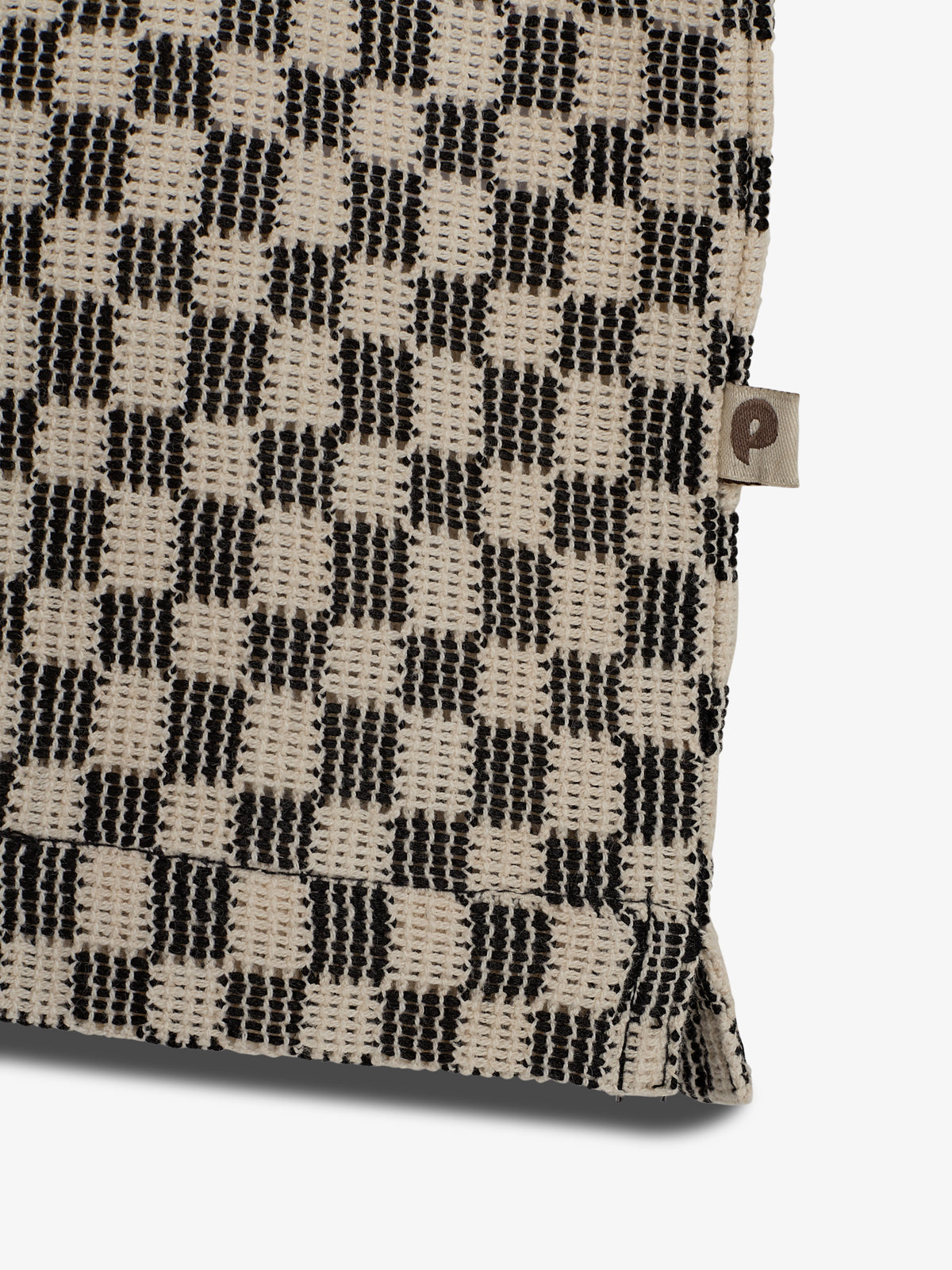 The object is a woven fabric with a checkered pattern of black and beige squares. A small tag featuring the letter "P" is attached. The background is plain white.