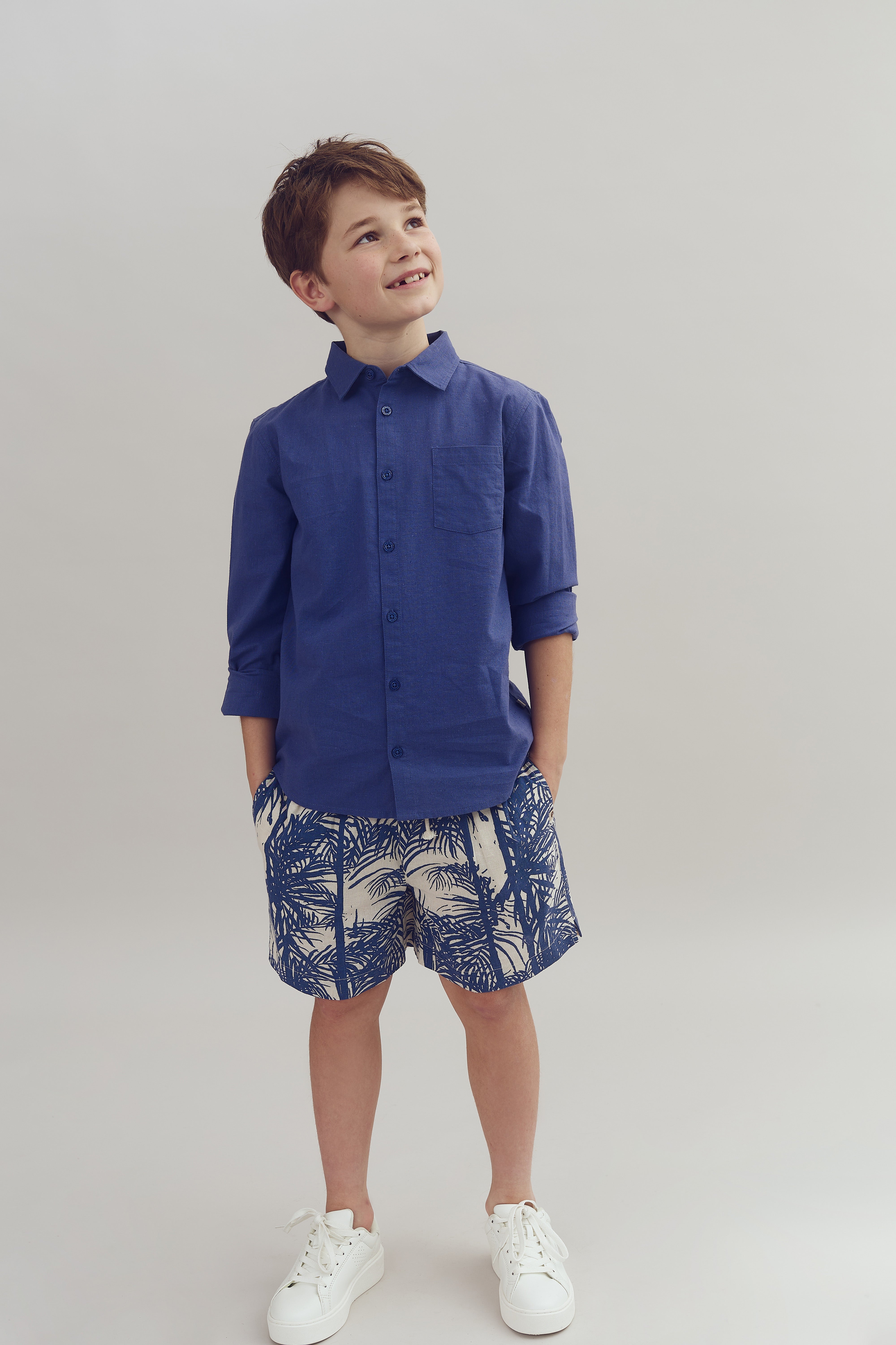 A smiling child stands with hands in pockets, wearing a blue shirt and patterned shorts, in a neutral indoor setting.