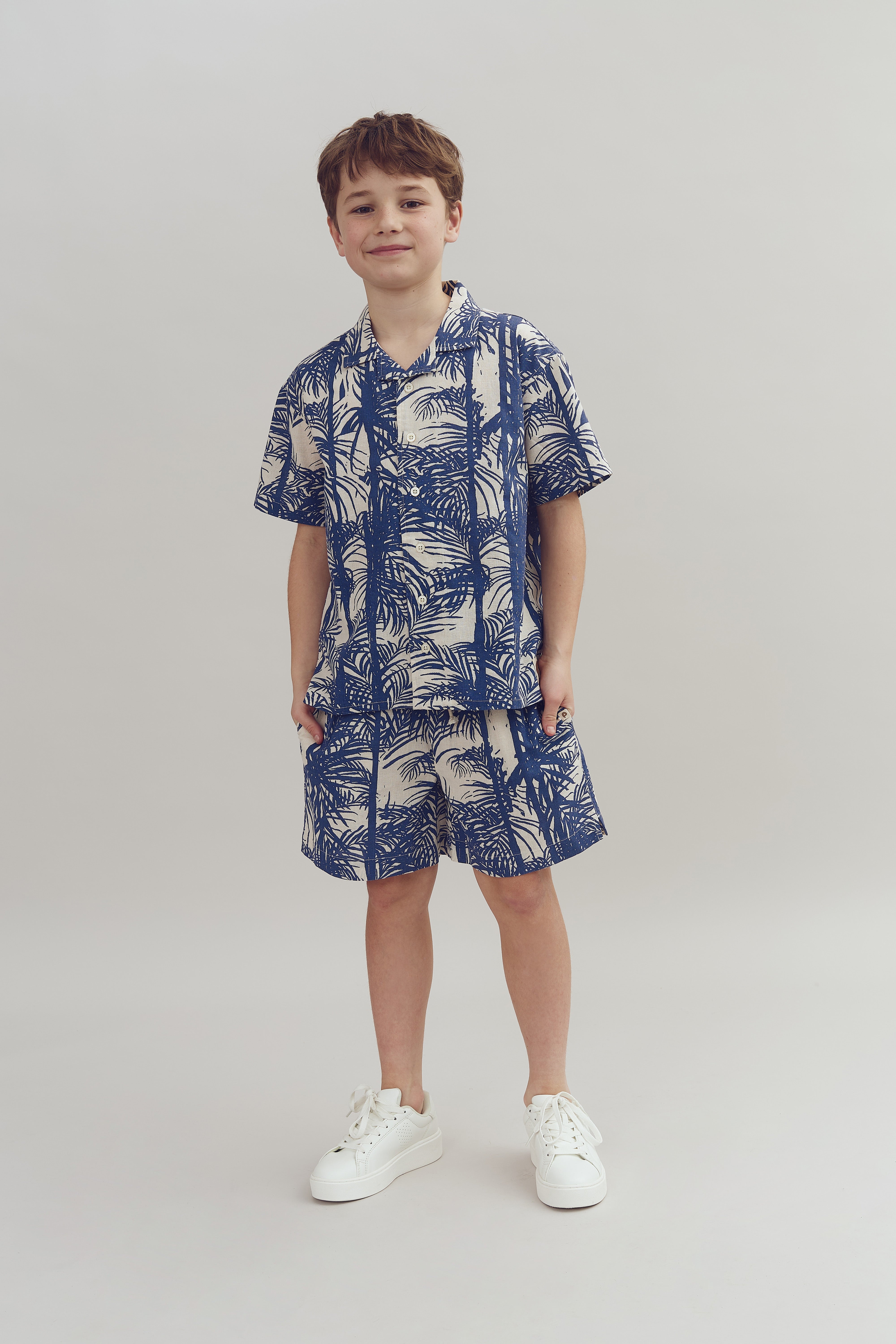 A young boy stands wearing a blue and white patterned shirt and shorts, paired with white sneakers. He is smiling slightly against a plain light-colored background.