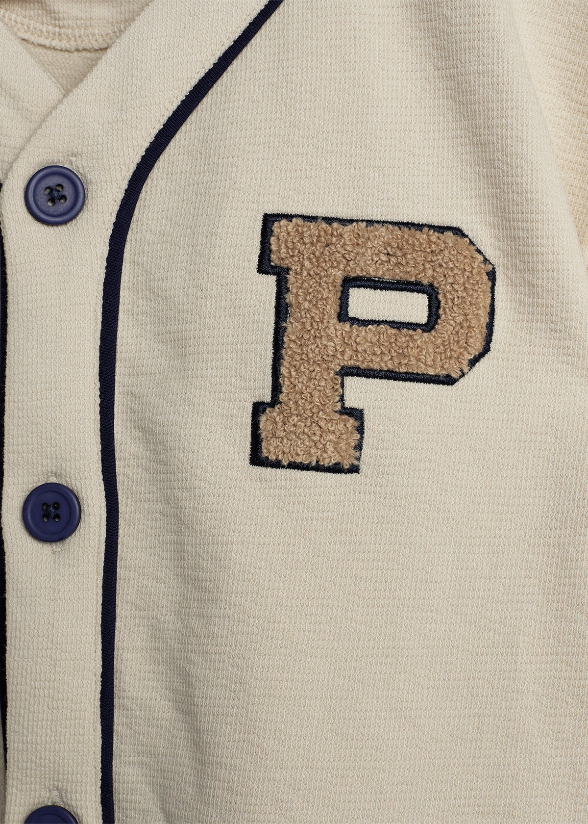 A beige fabric features a large textured letter "P" embroidered in brown. Blue buttons and dark trim line the garment's edges, suggesting a sporty jacket or jersey in a casual setting.