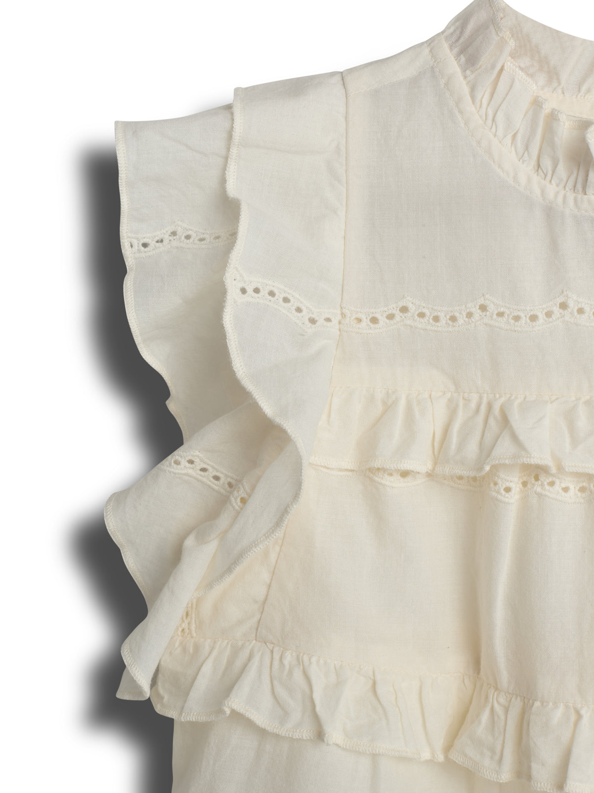 A cream-colored blouse features ruffled layers and lace details. It appears in a close-up shot, showcasing the intricate texture and design, with soft shadows cast in the surrounding area.