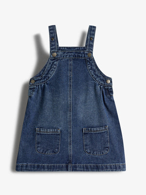 Denim pinafore dress hangs against a plain white background, featuring adjustable straps with buttons and two front pockets. The dress has a simple, casual design.
