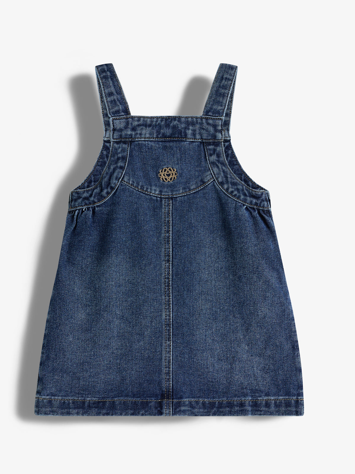 Denim pinafore dress with shoulder straps, featuring a decorative emblem on the front. The backdrop is a plain white surface.