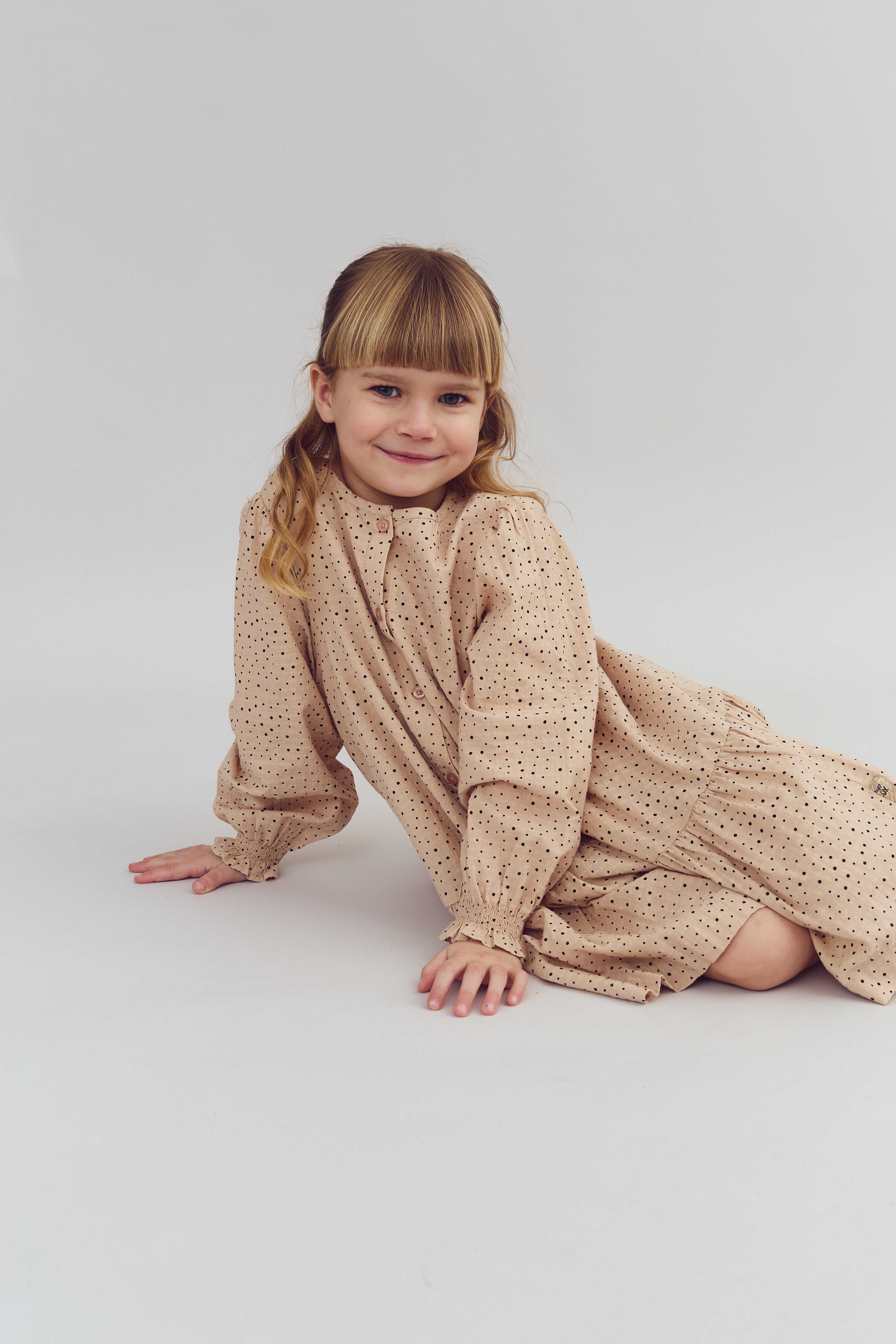 A young girl sits and smiles, wearing a beige polka-dot dress in a minimal studio setting with a neutral background.