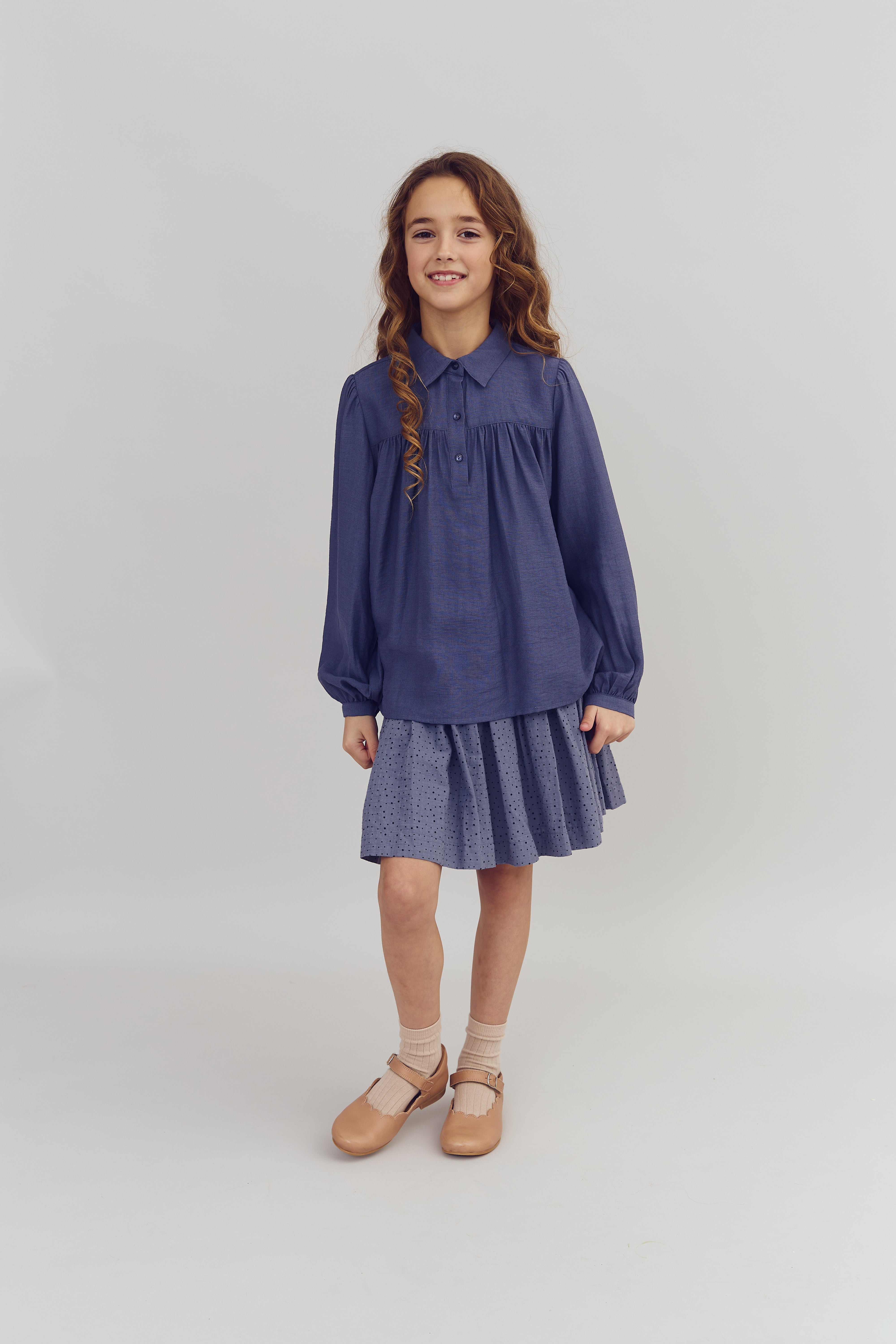 A young girl stands smiling, wearing a blue blouse and skirt, with beige shoes and socks. She has wavy, long hair. The background is a plain, light-colored studio setting.