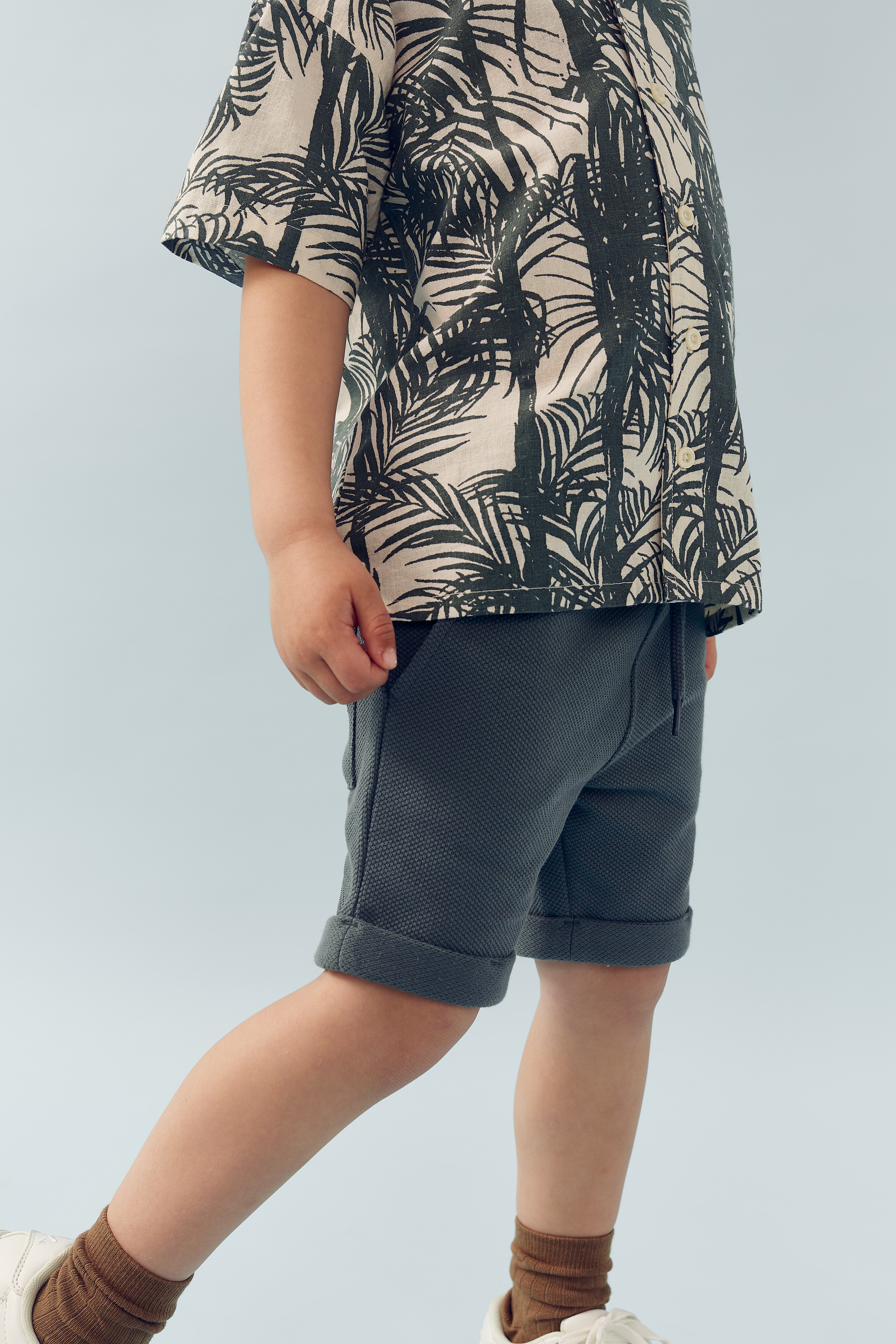 Child wearing a palm-patterned shirt and gray shorts stands in a neutral environment. Their feet are in white sneakers and brown socks. The background is a light blue shade.