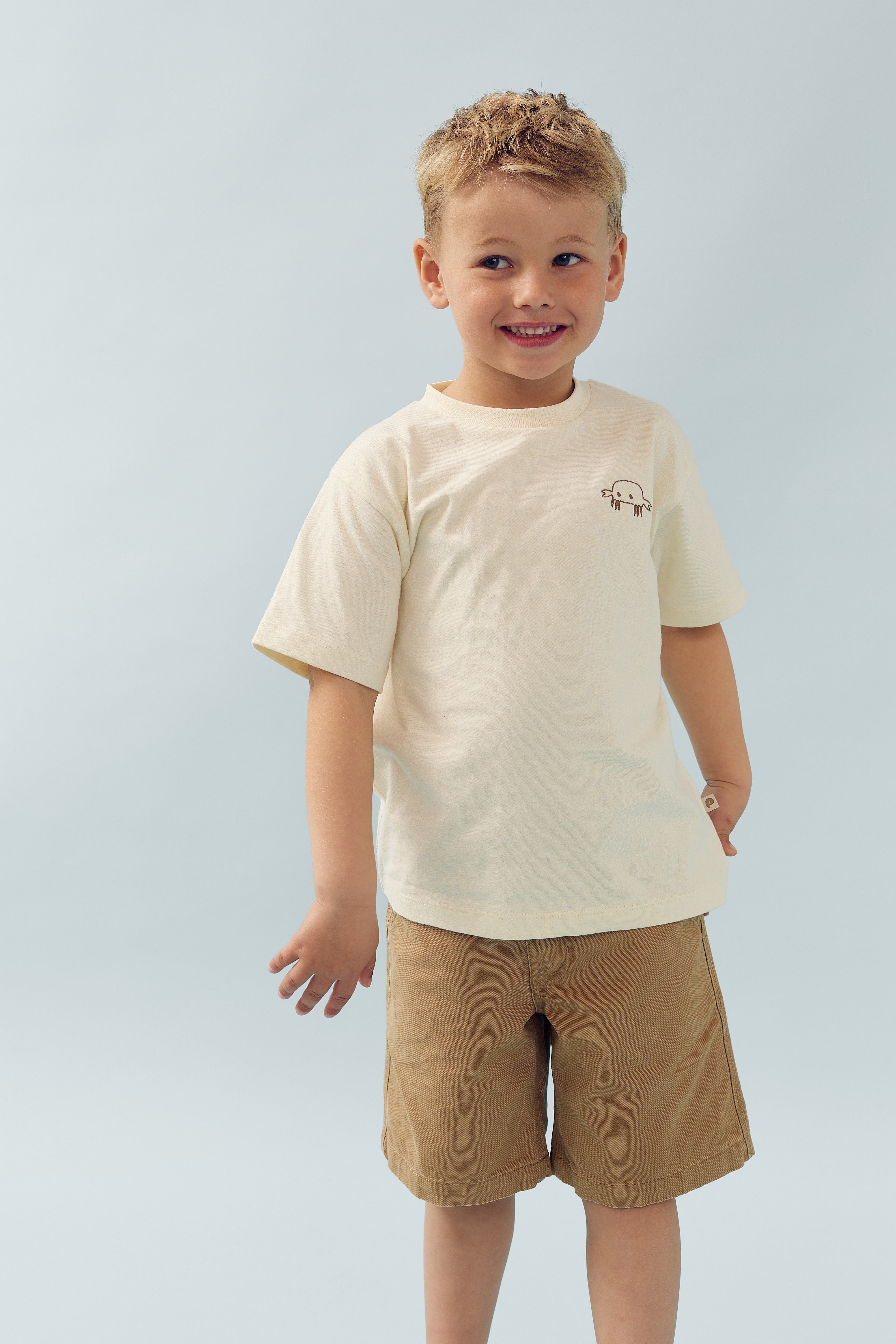 A smiling child stands against a plain light blue background, wearing a cream t-shirt with a small crab graphic, and brown shorts.