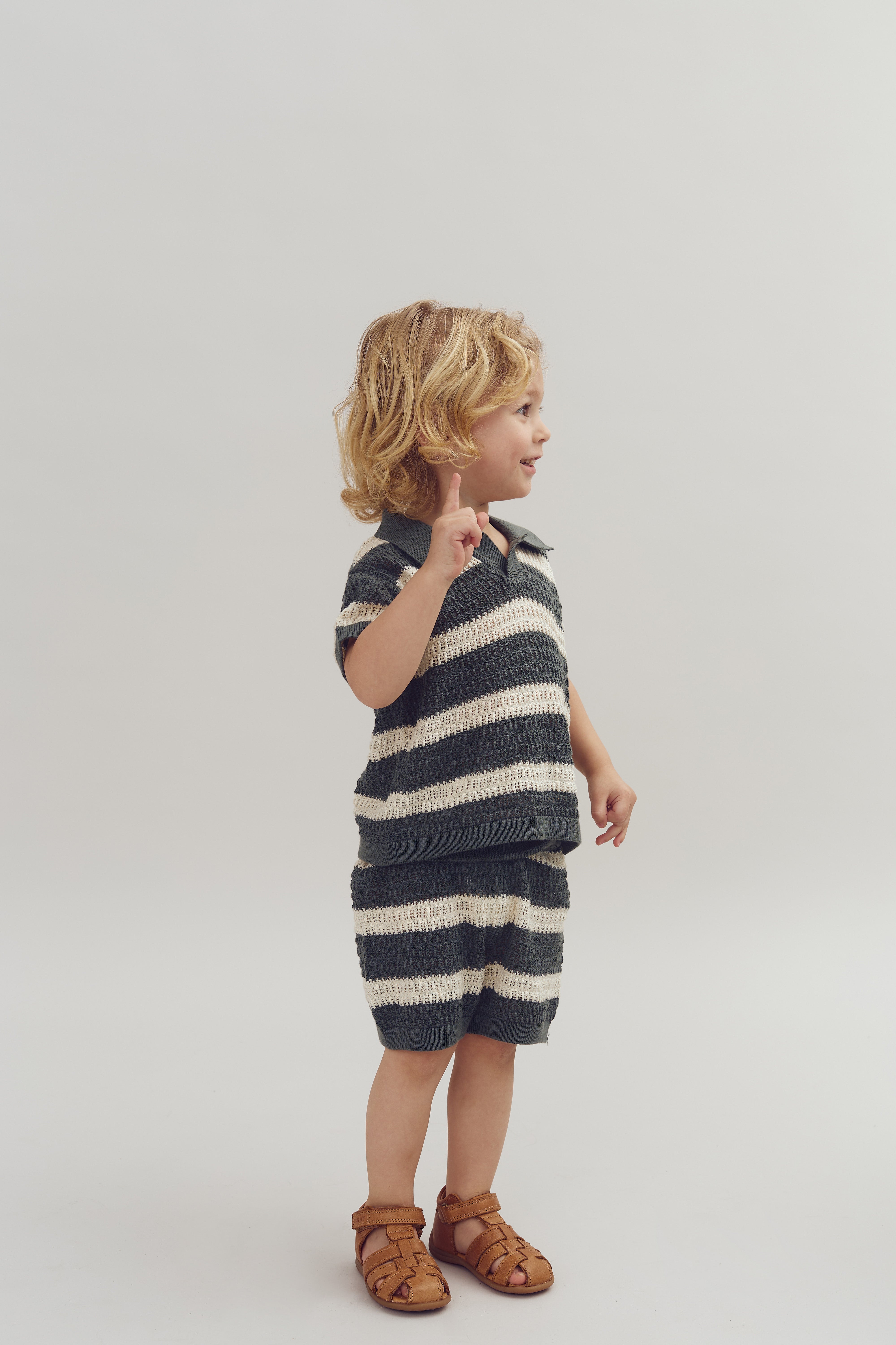 Child wearing striped knit outfit stands, pointing up with finger, in a studio setting with a plain light background. Brown sandals complete the look.