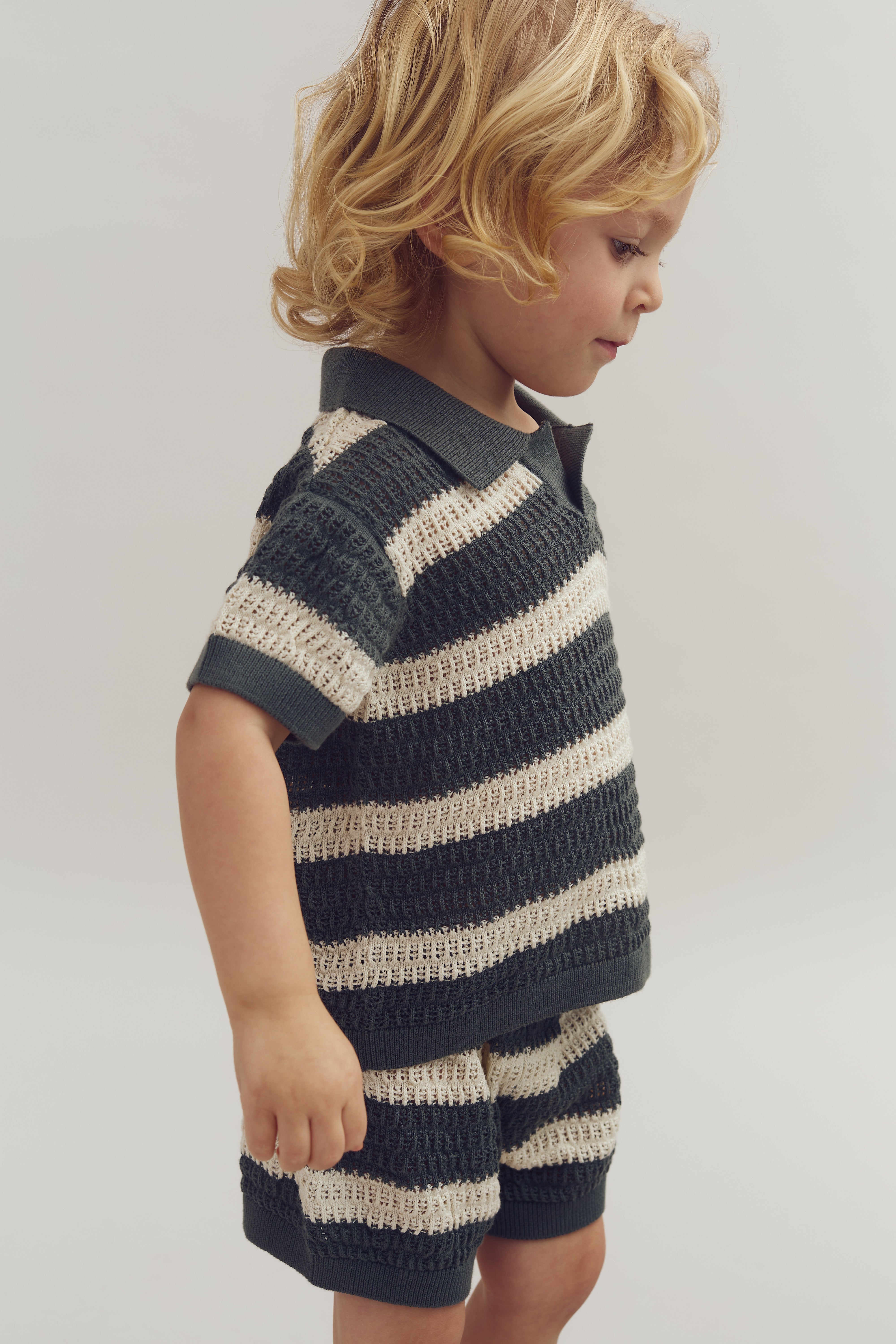 Child standing, wearing a black and white striped knit outfit with shorts, looking to the side. The background is a plain, light-colored setting.