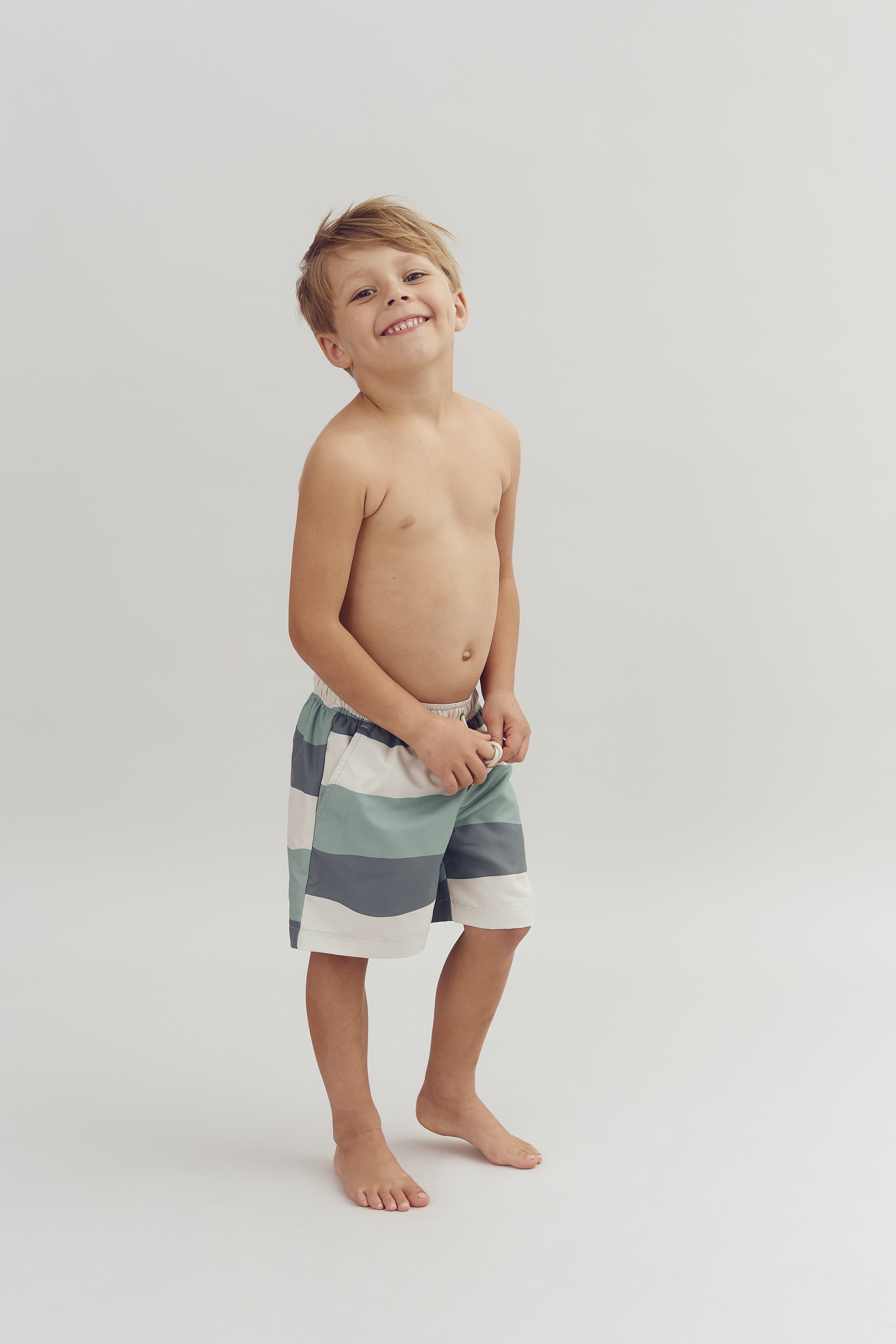 A child, smiling broadly, stands barefoot wearing striped shorts against a plain white background, exuding a playful and carefree demeanor.