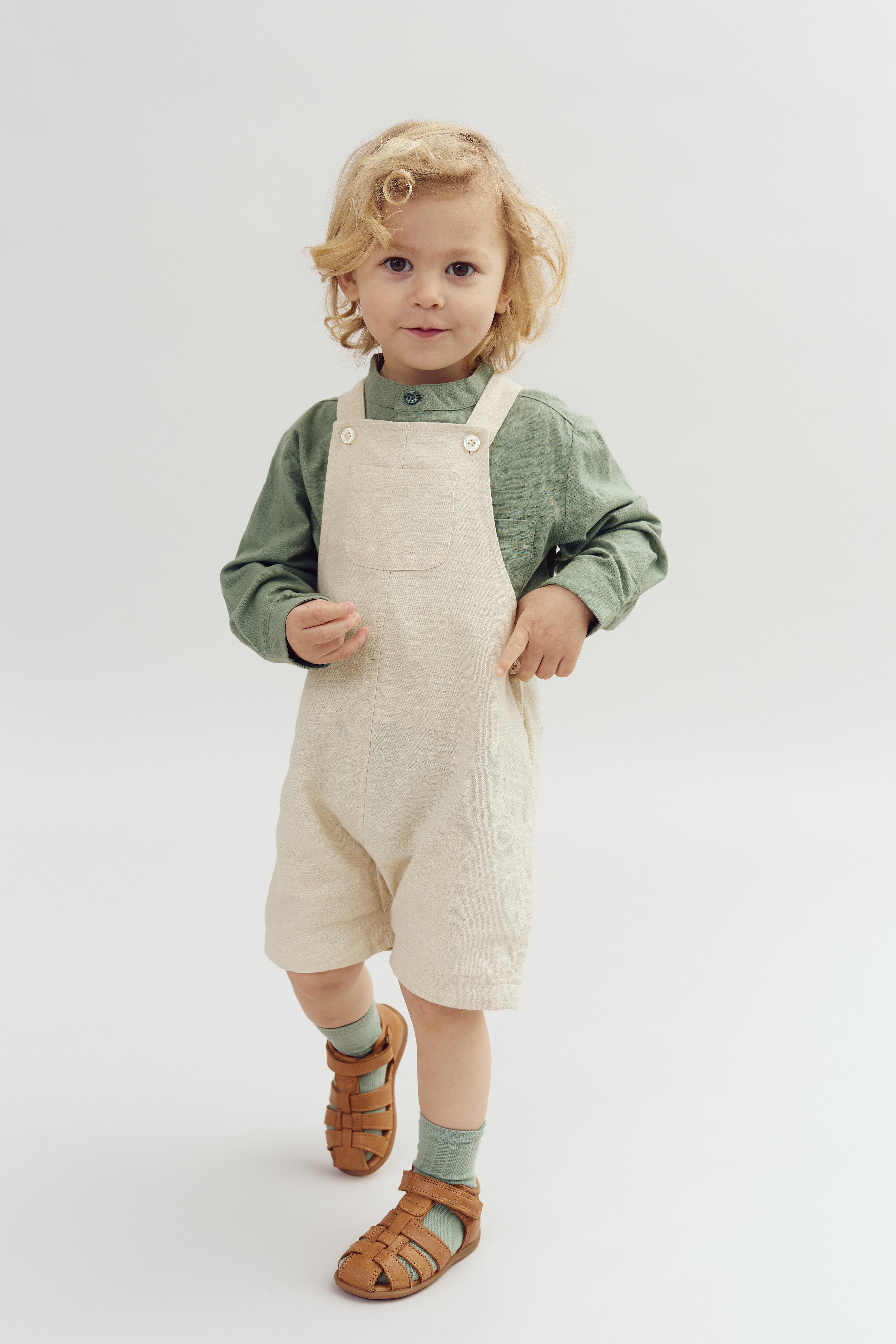 A young child with curly blonde hair stands wearing a green long-sleeve shirt, beige overalls, green socks, and brown sandals against a plain white background, gazing calmly forward.