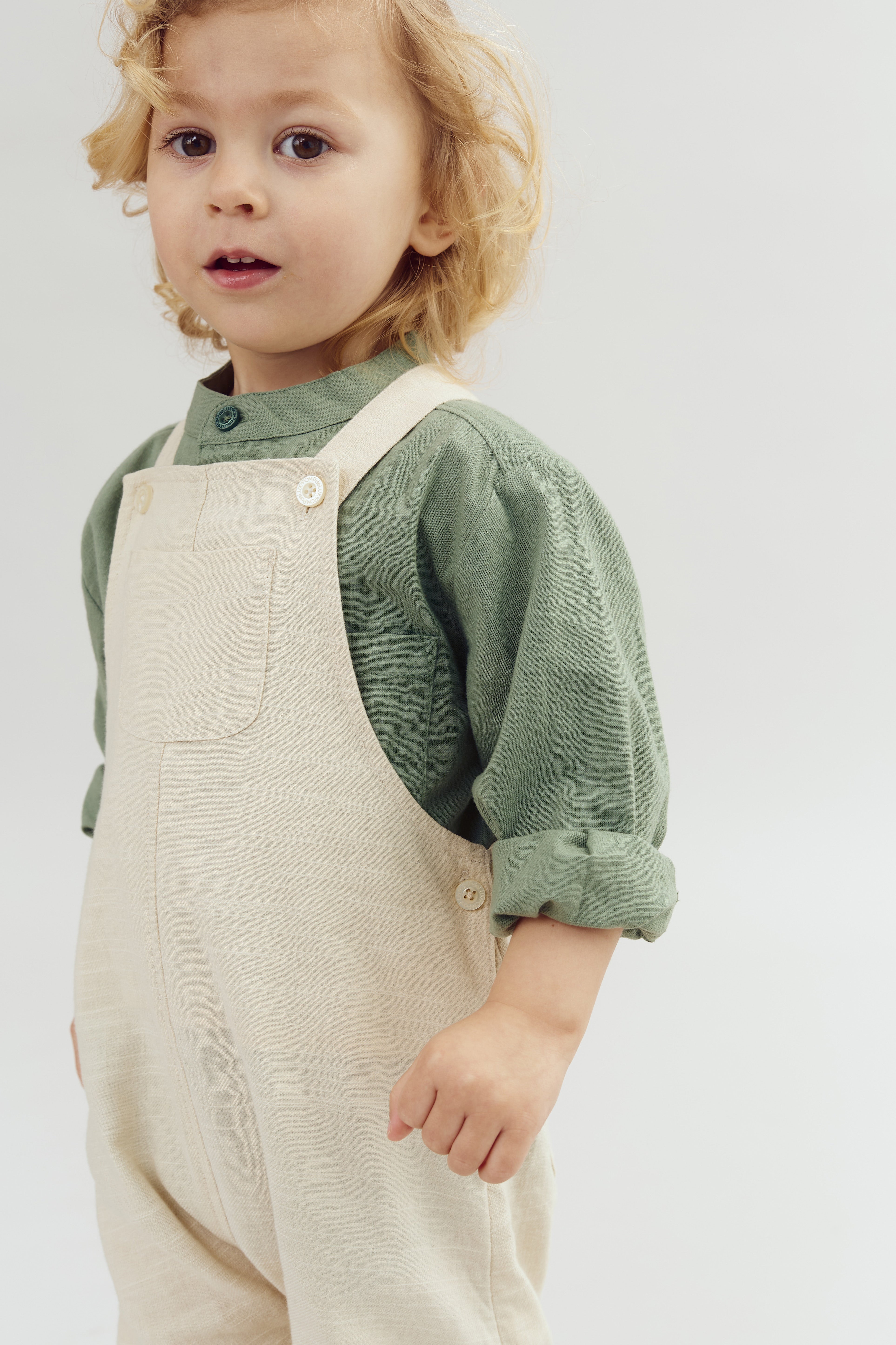 A young child with curly blonde hair wears a beige overall and green shirt, looking ahead with a neutral expression, against a plain, light background.