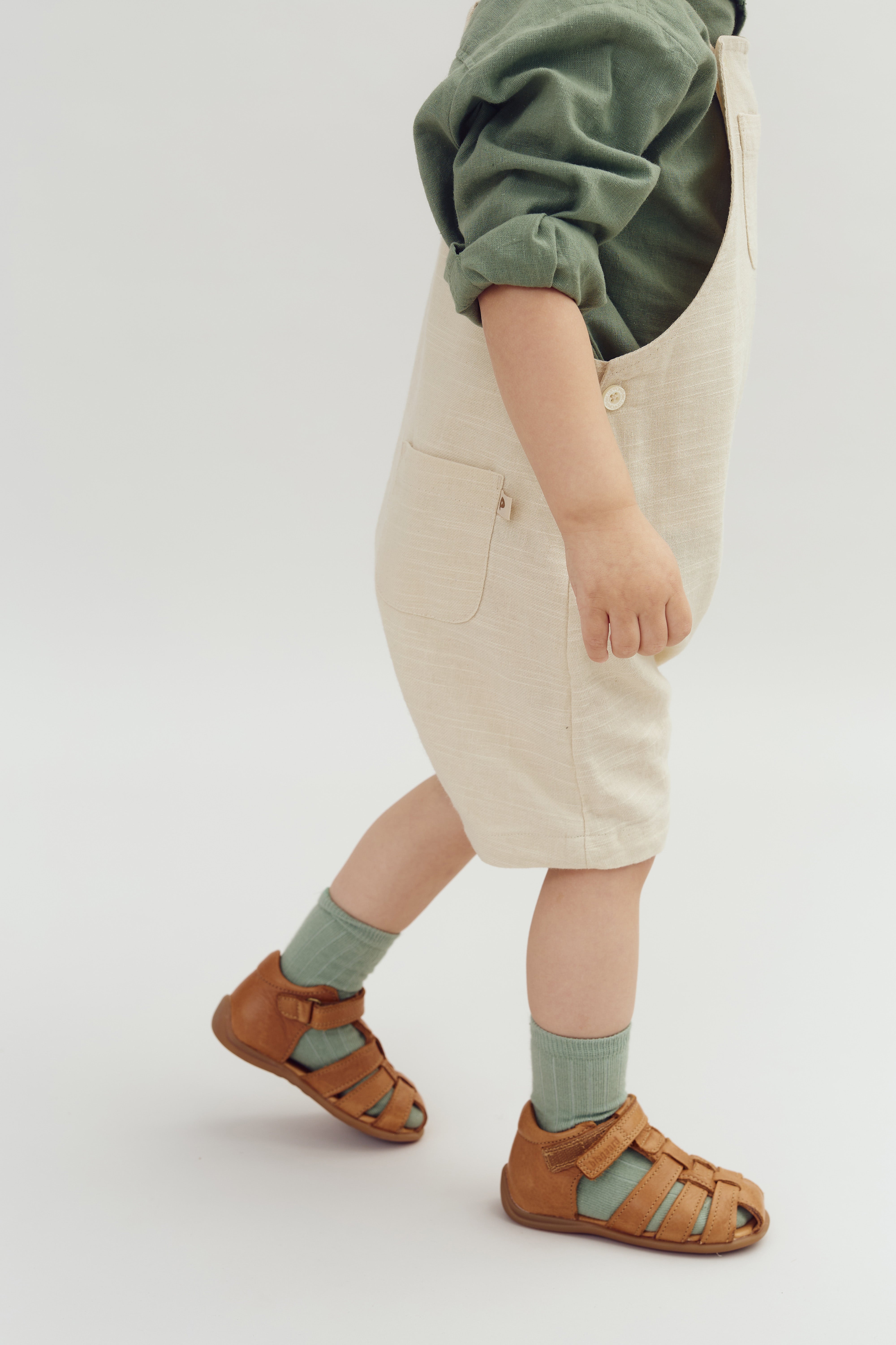 Child wearing tan overalls, green socks, and brown sandals walks forward. The setting is a plain, neutral background.