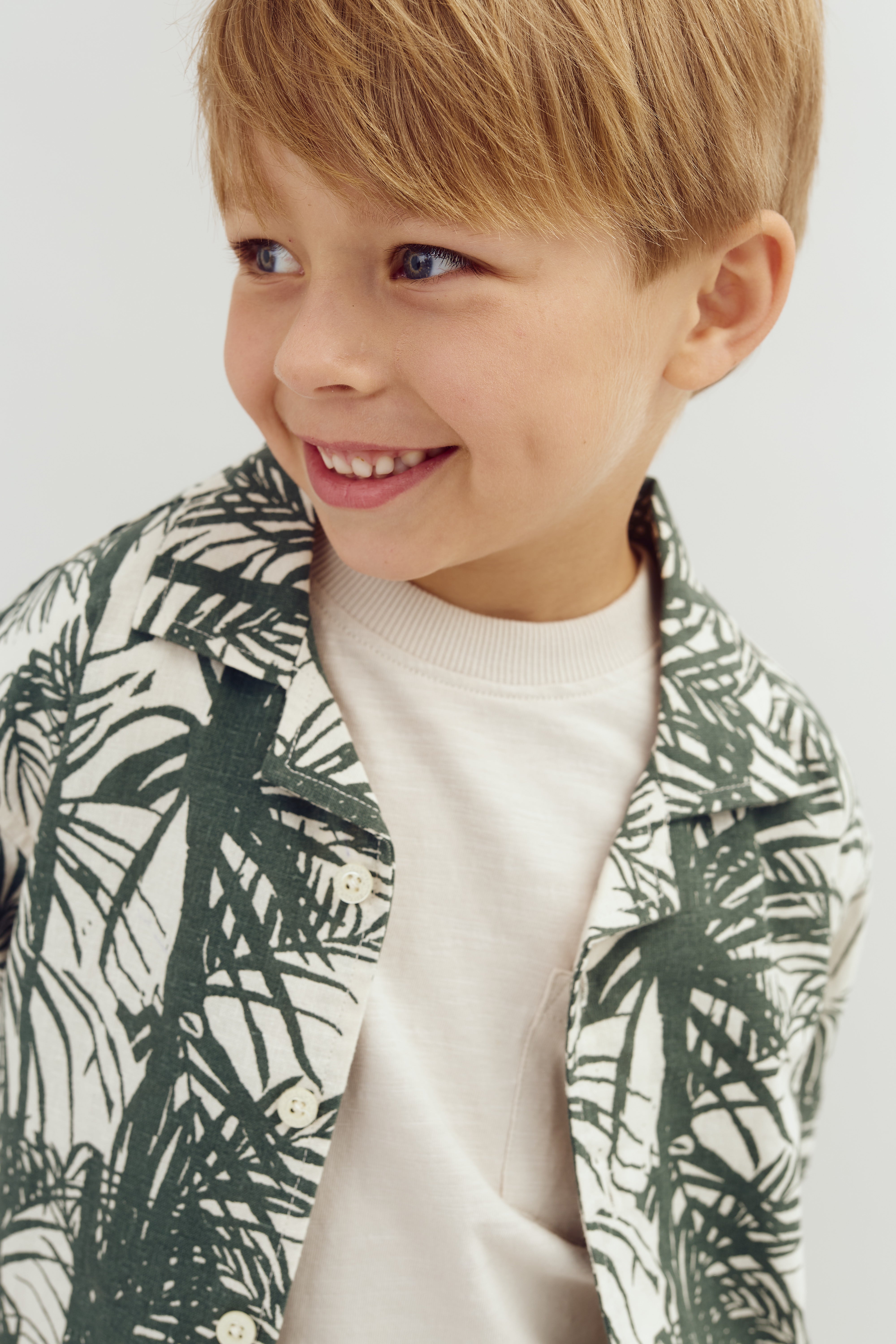 A smiling child in a leaf-patterned shirt looks to the side against a plain white background.
