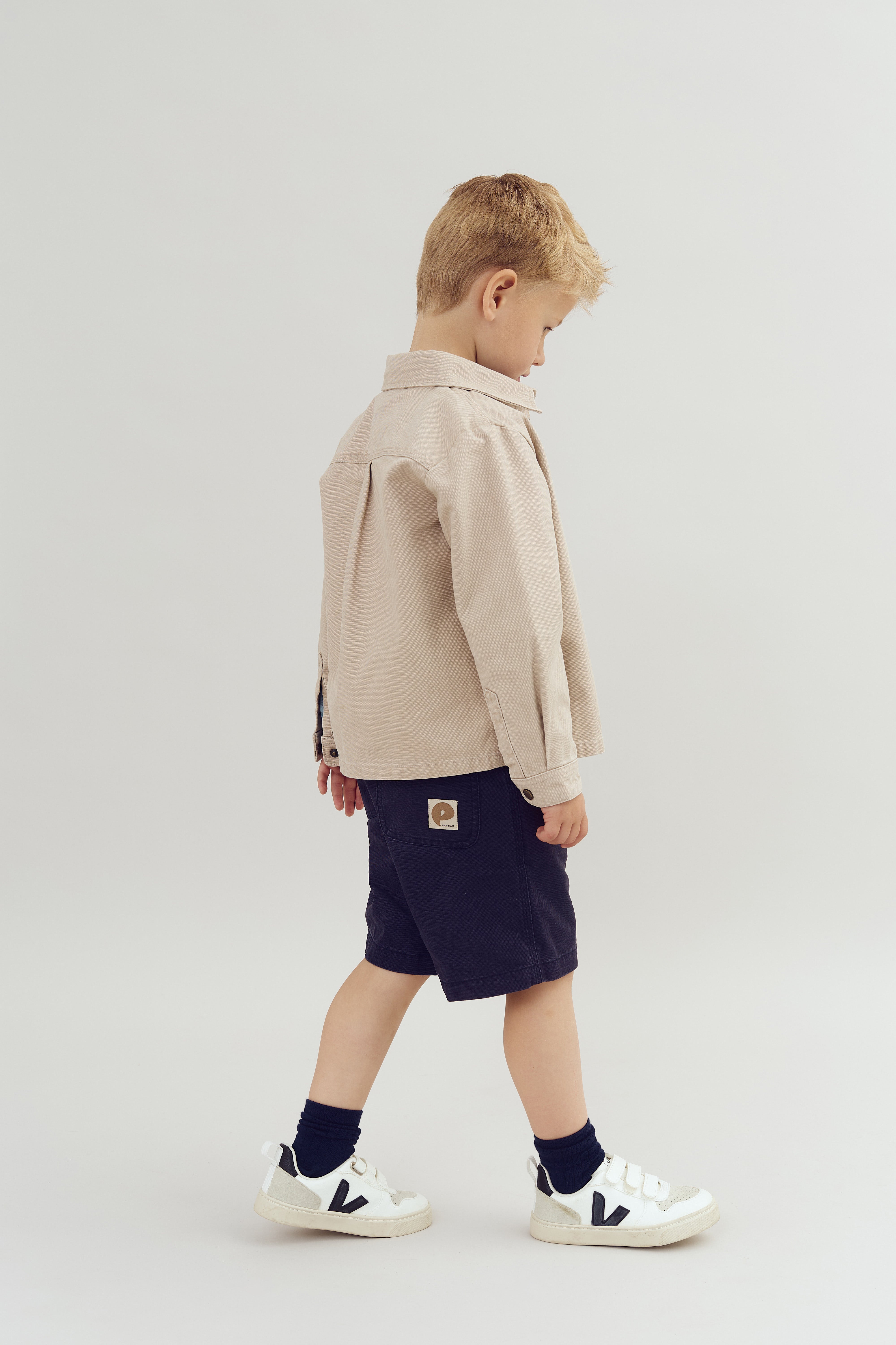 A child, wearing a beige shirt and dark shorts with a patch, walks forward. The shoes are white with black accents. The background is plain and light-colored.