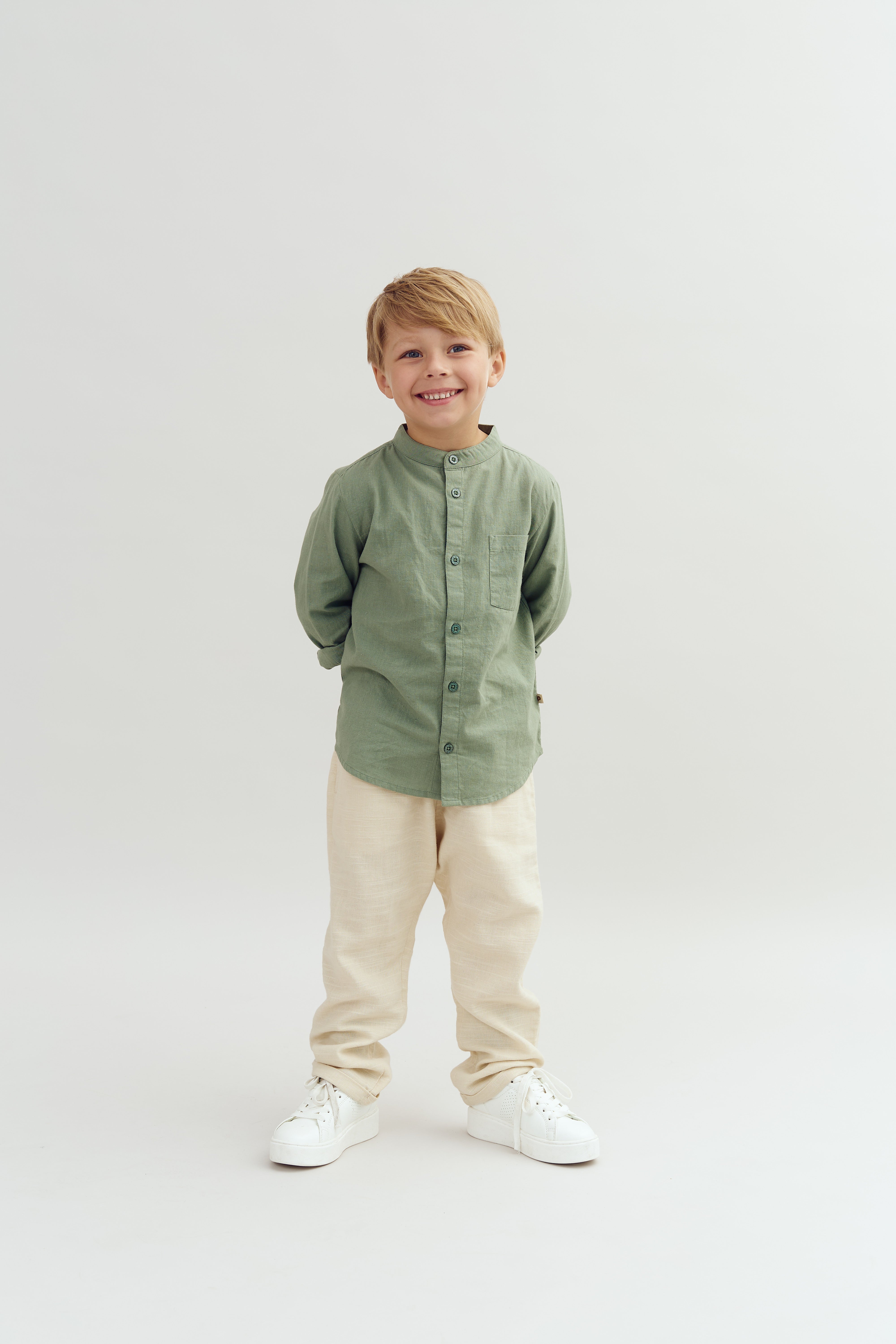 A young child, smiling with hands behind their back, wears a green shirt, beige pants, and white sneakers, standing against a plain, light gray background.