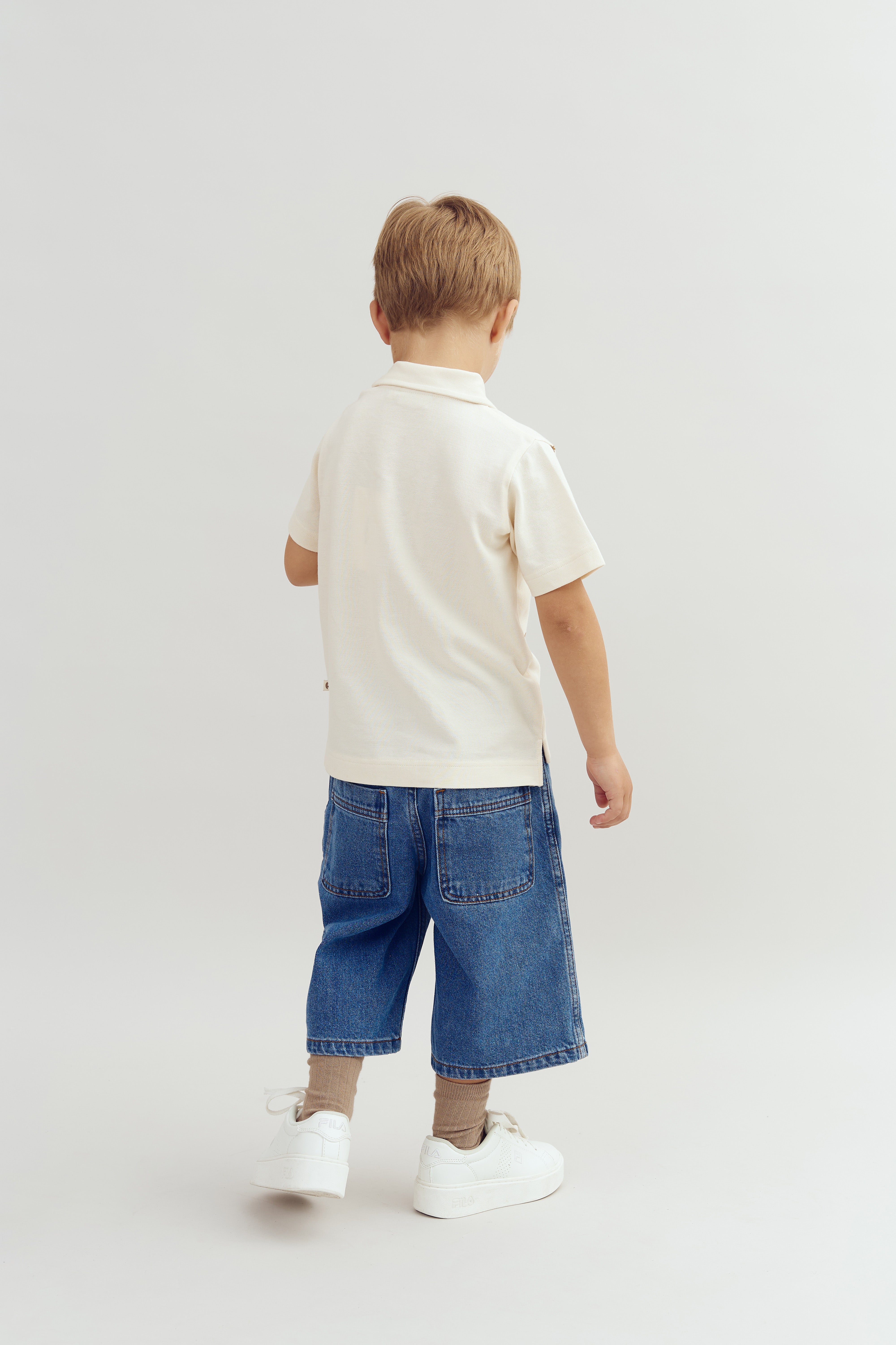 A child stands facing away, wearing a white shirt, denim shorts, and white shoes, in a minimalist, light-colored studio setting.