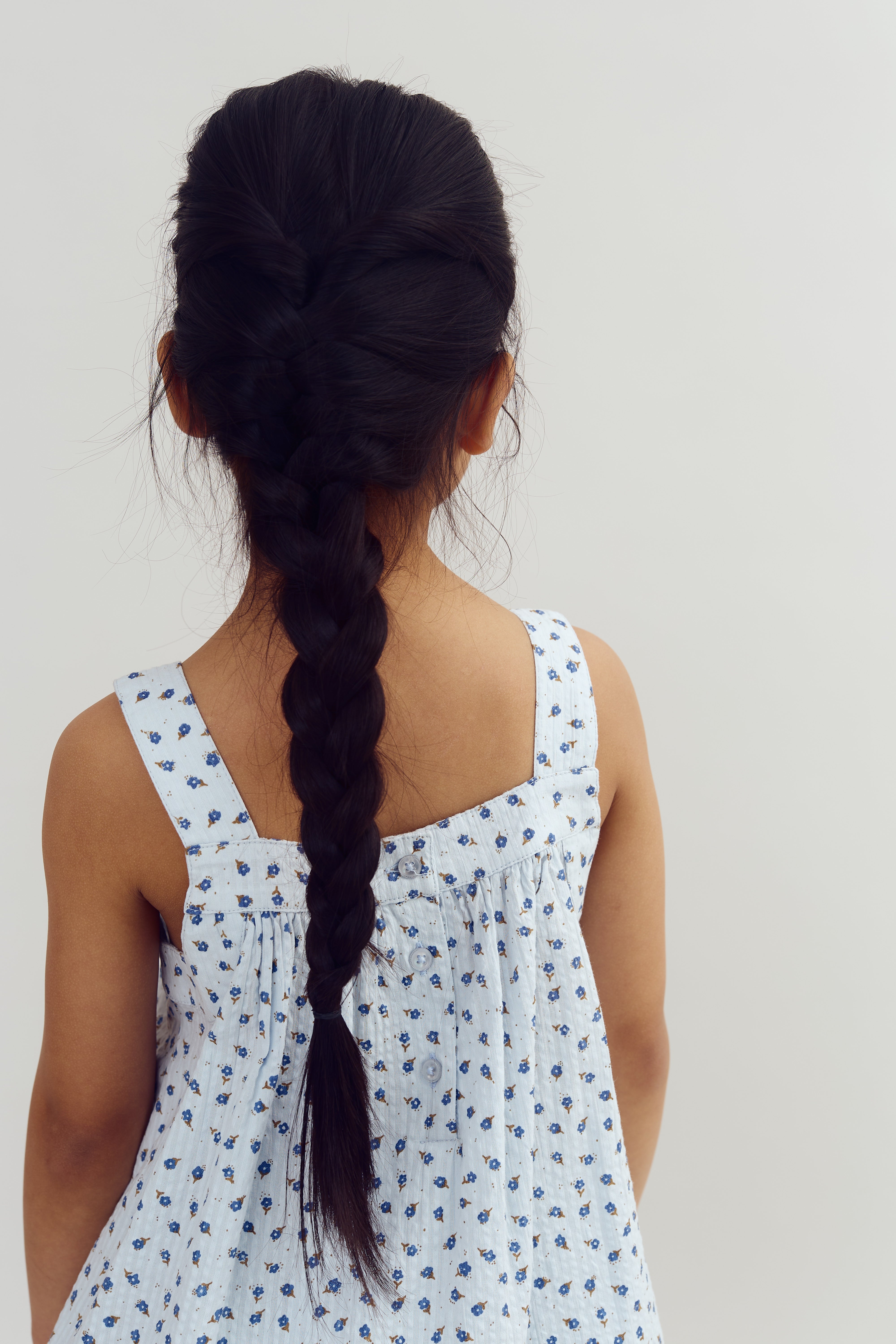 A child with long, dark braided hair stands facing away, wearing a light blue dress adorned with small blue and brown floral patterns. The background is plain white.
