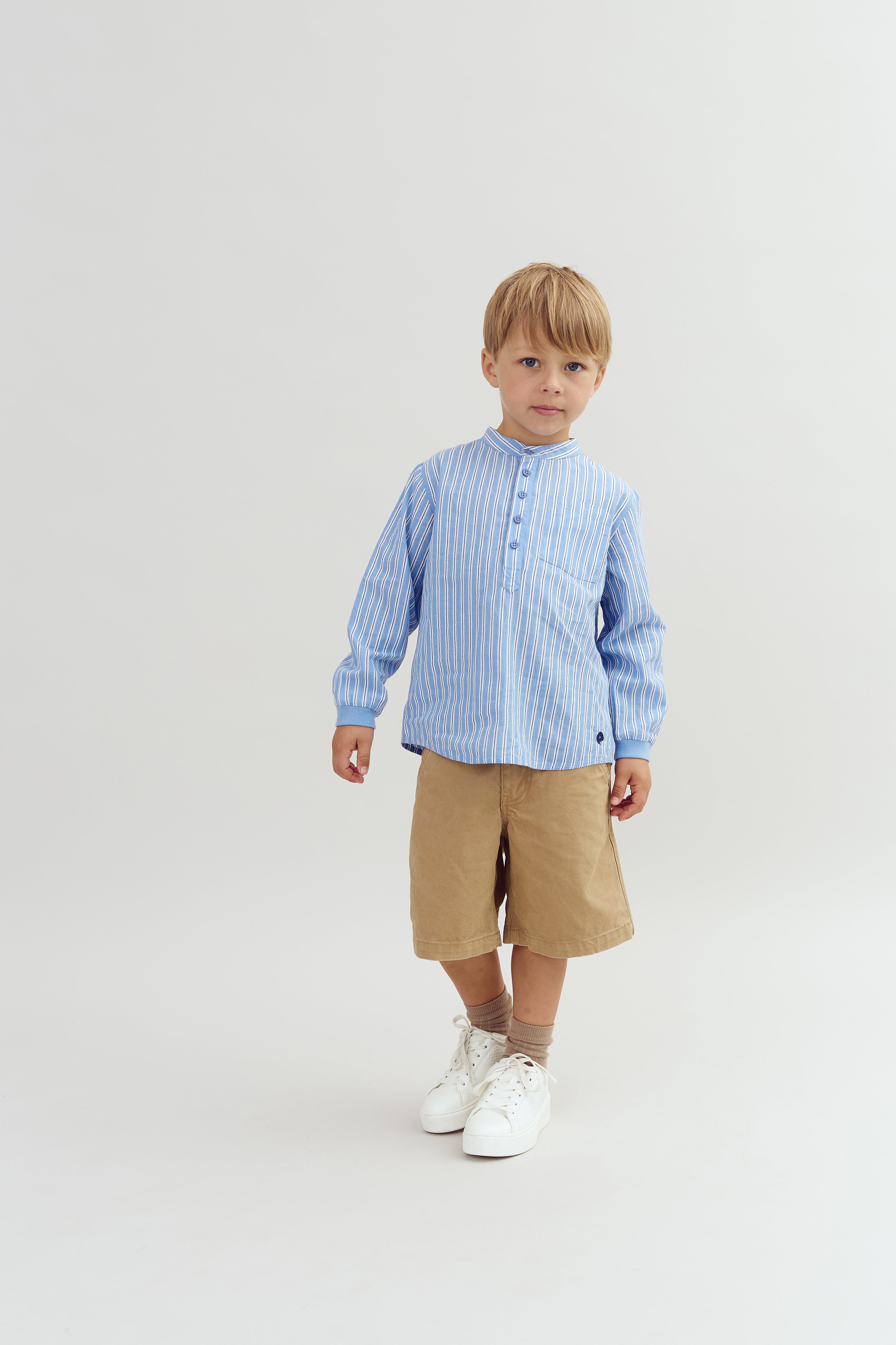 A child stands in a light blue striped shirt and tan shorts against a plain white background, looking directly forward with a neutral expression.
