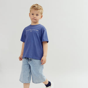 A child in a blue shirt is walking, with text "Don’t be crabby, just be happy" on the shirt, wearing denim shorts and sneakers, against a plain background.