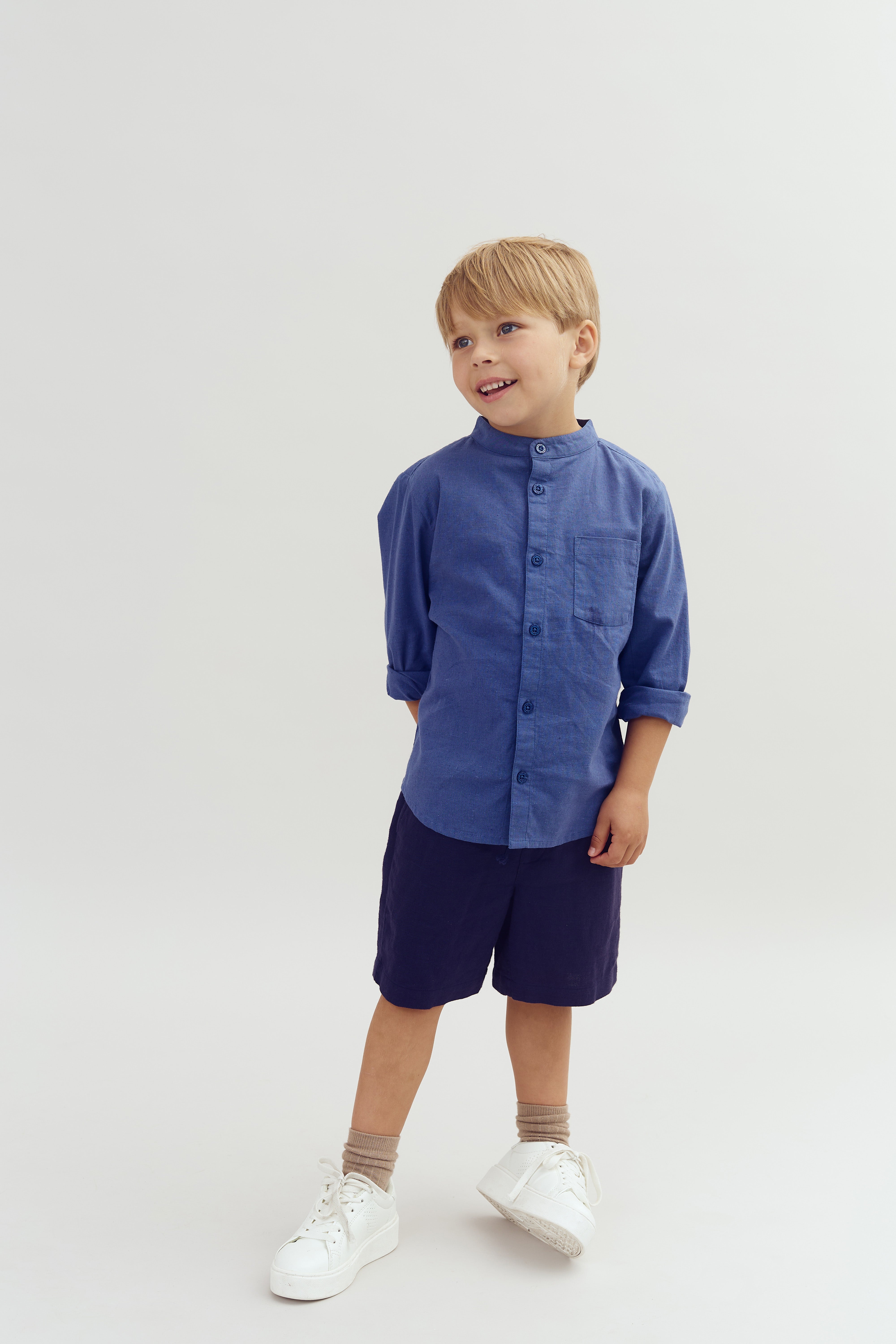 A young child stands smiling, hands behind the back, wearing a blue shirt and dark shorts. The child has light hair, with the background plain and light-colored.