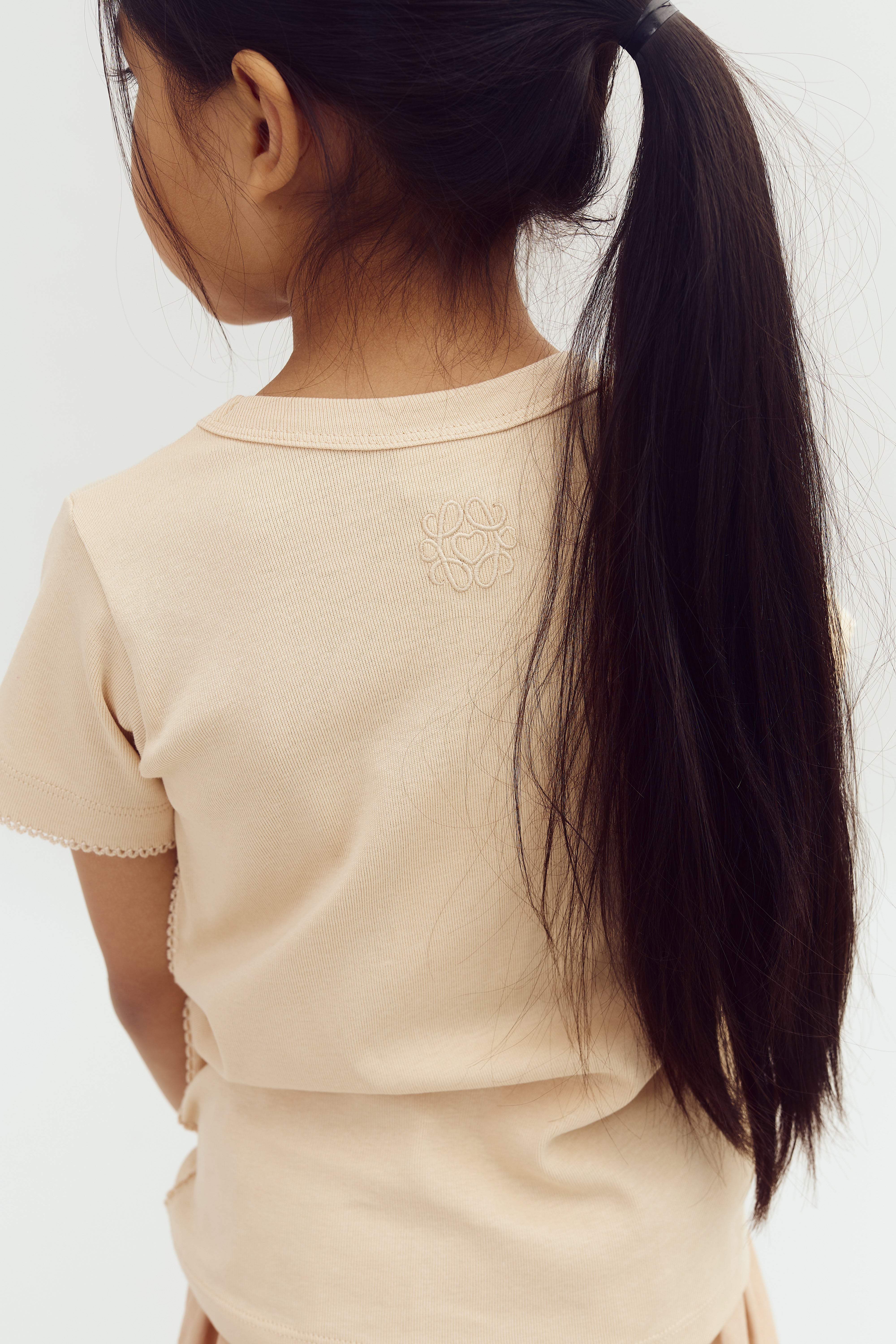 A child with long brown hair in a ponytail, wearing a beige shirt with intricate embroidery on the back, stands against a plain white background.