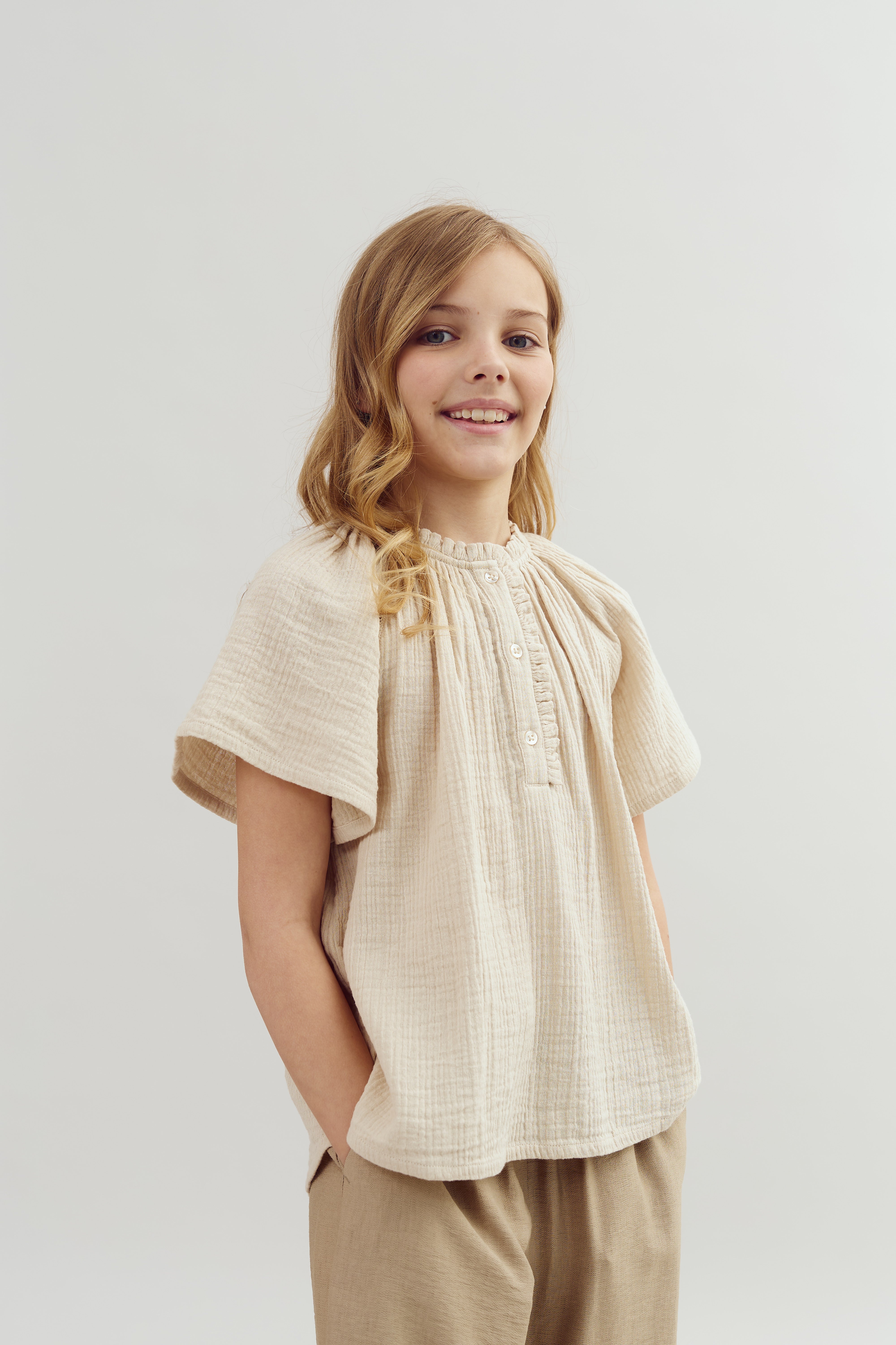 A young person stands smiling, wearing a cream-colored blouse and beige pants, with hands in pockets against a plain white background.