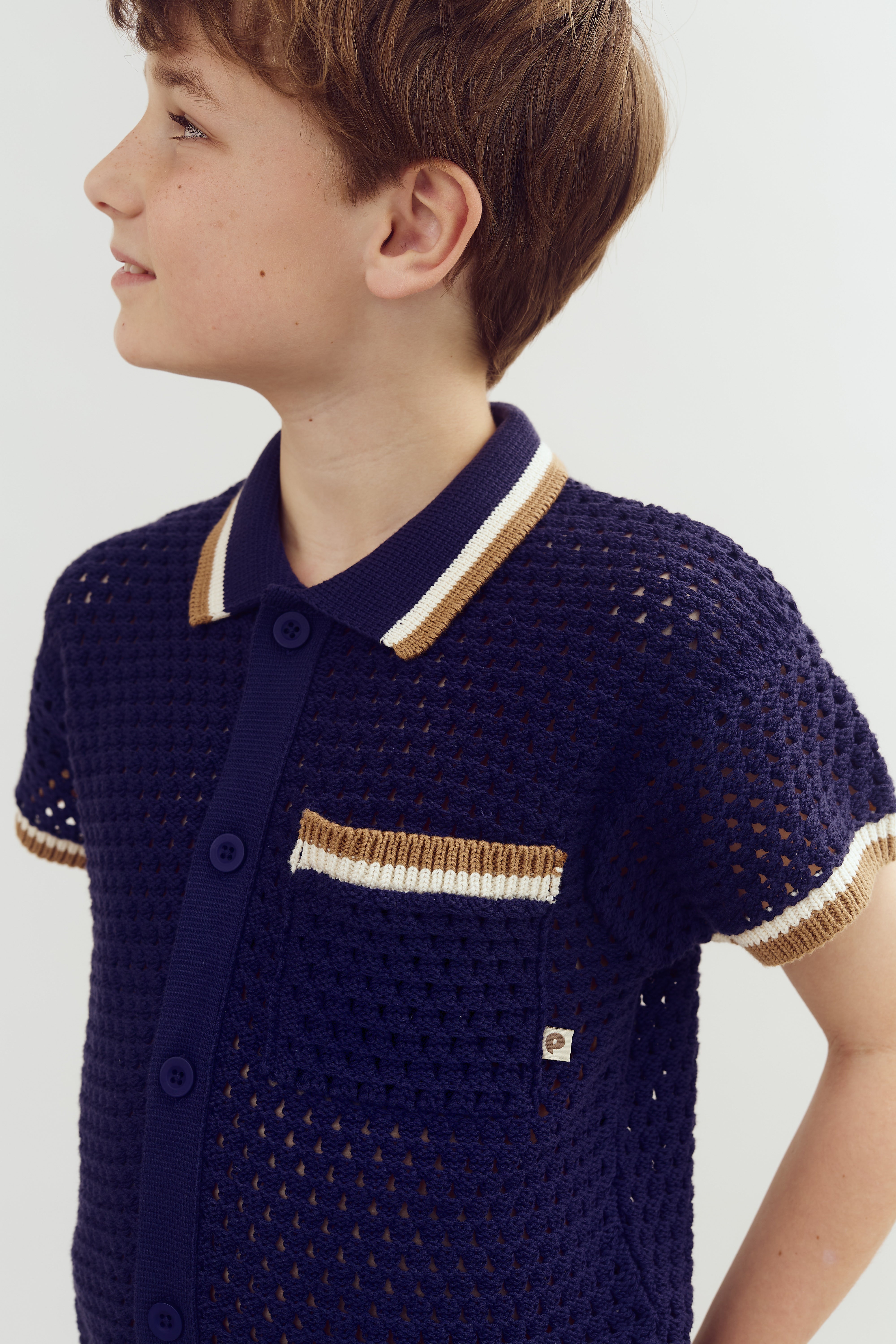 A person models a navy blue crochet shirt with beige and white trim, featuring a pocket and buttons, against a plain white background.